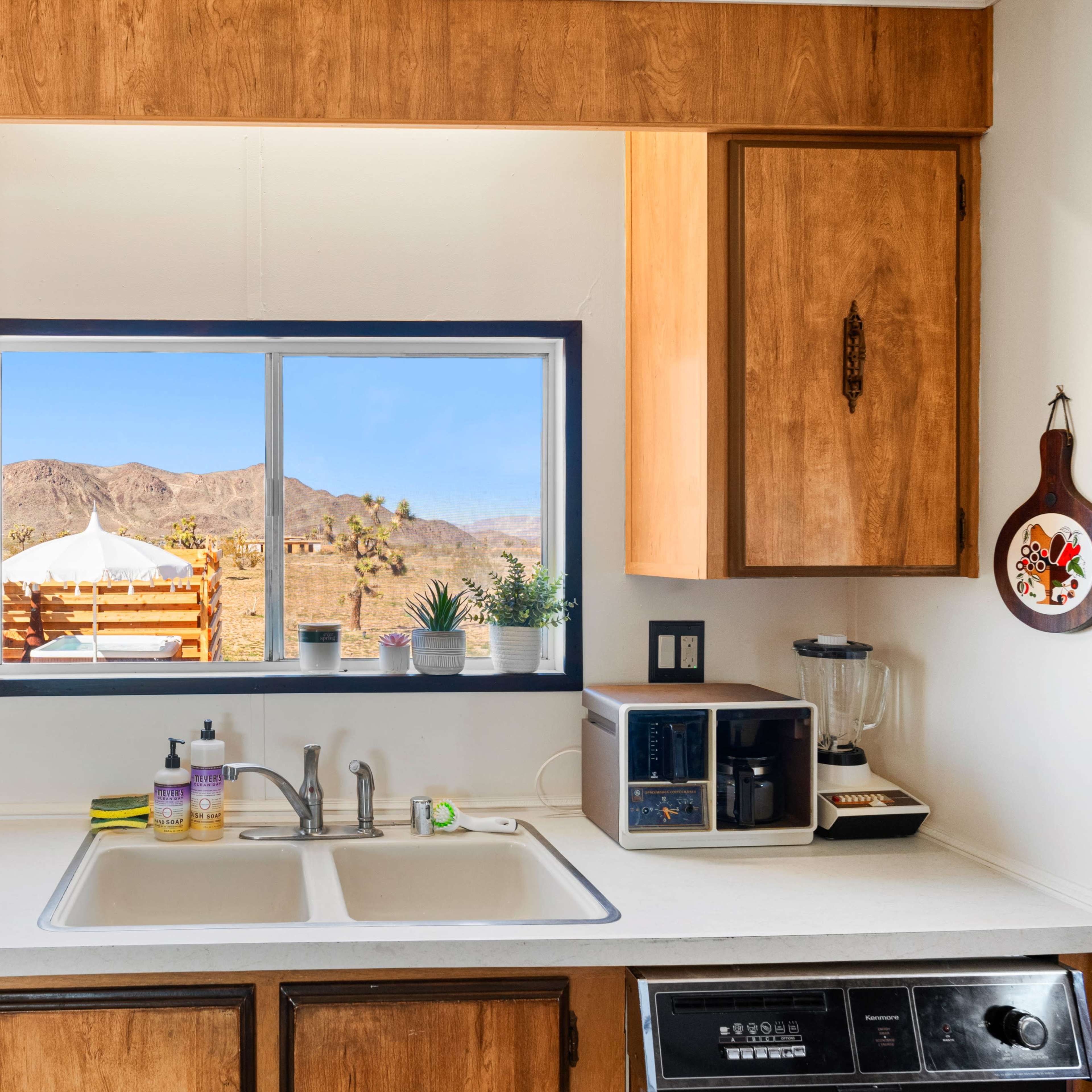 A kitchen with wooden cabinets and a clean countertop features a window displaying a desert landscape and outdoor seating.