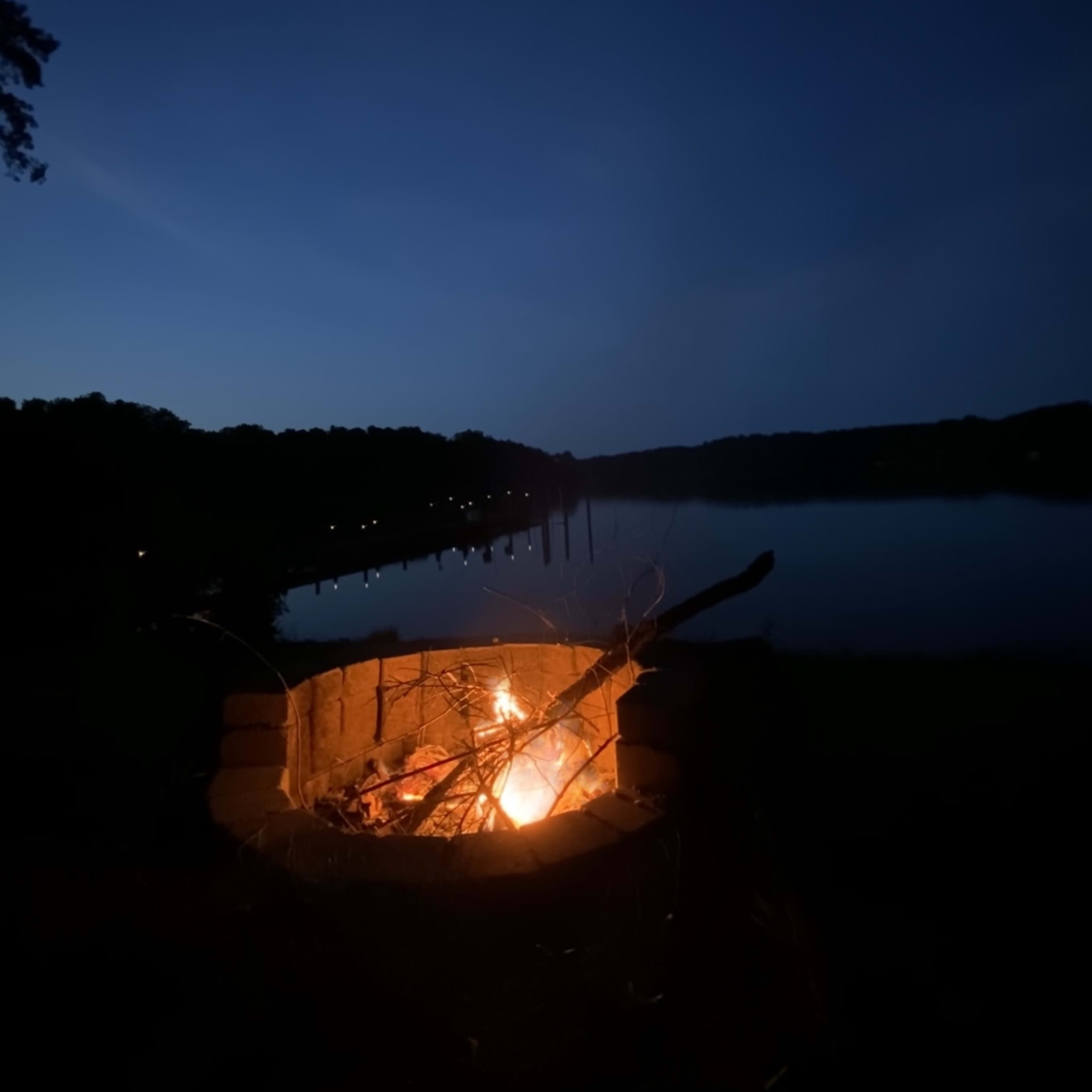 A fire burns in a stone pit beside a calm lake under a twilight sky.