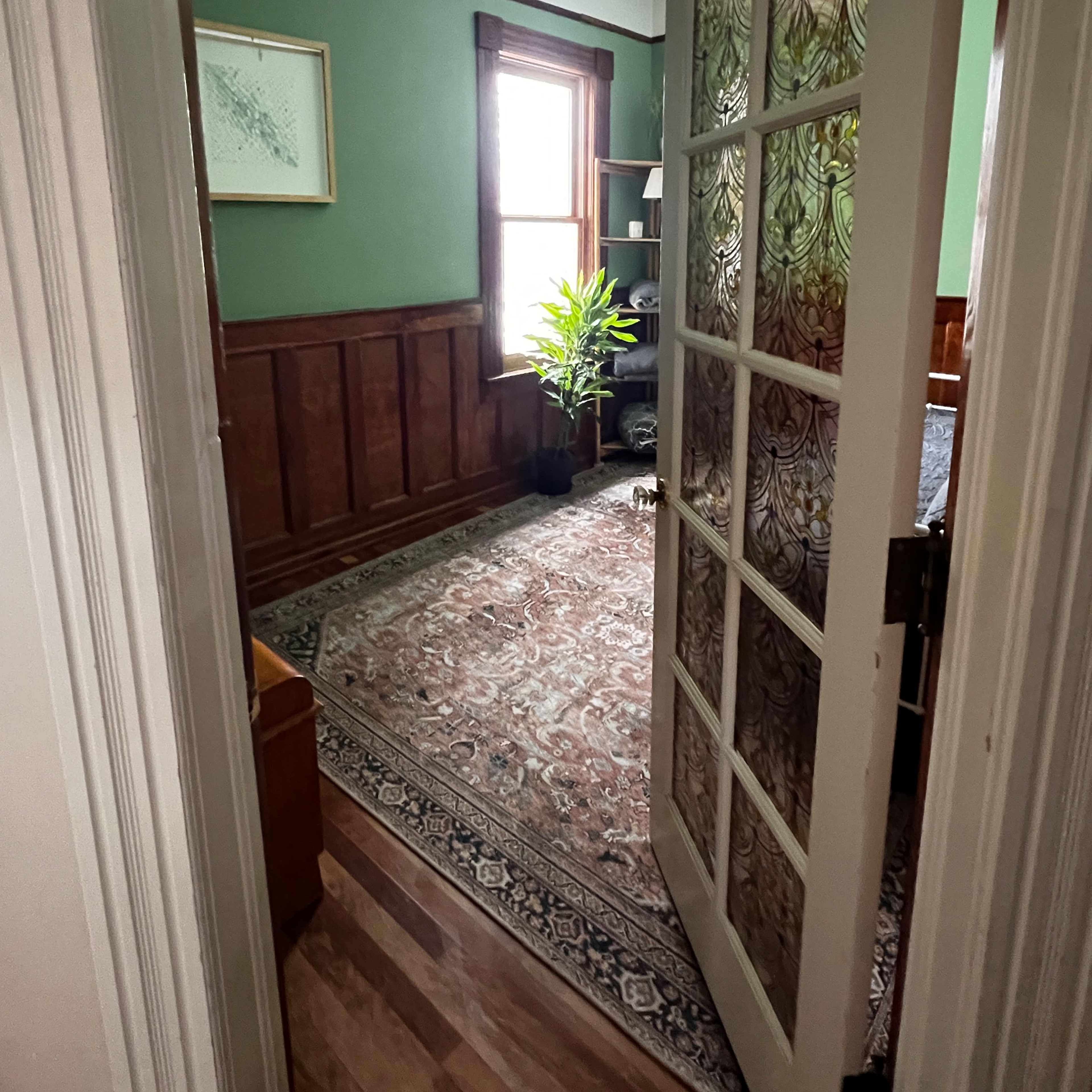 A partially open door reveals a room with green walls, a wooden floor, a potted plant by a window, and a patterned rug.
