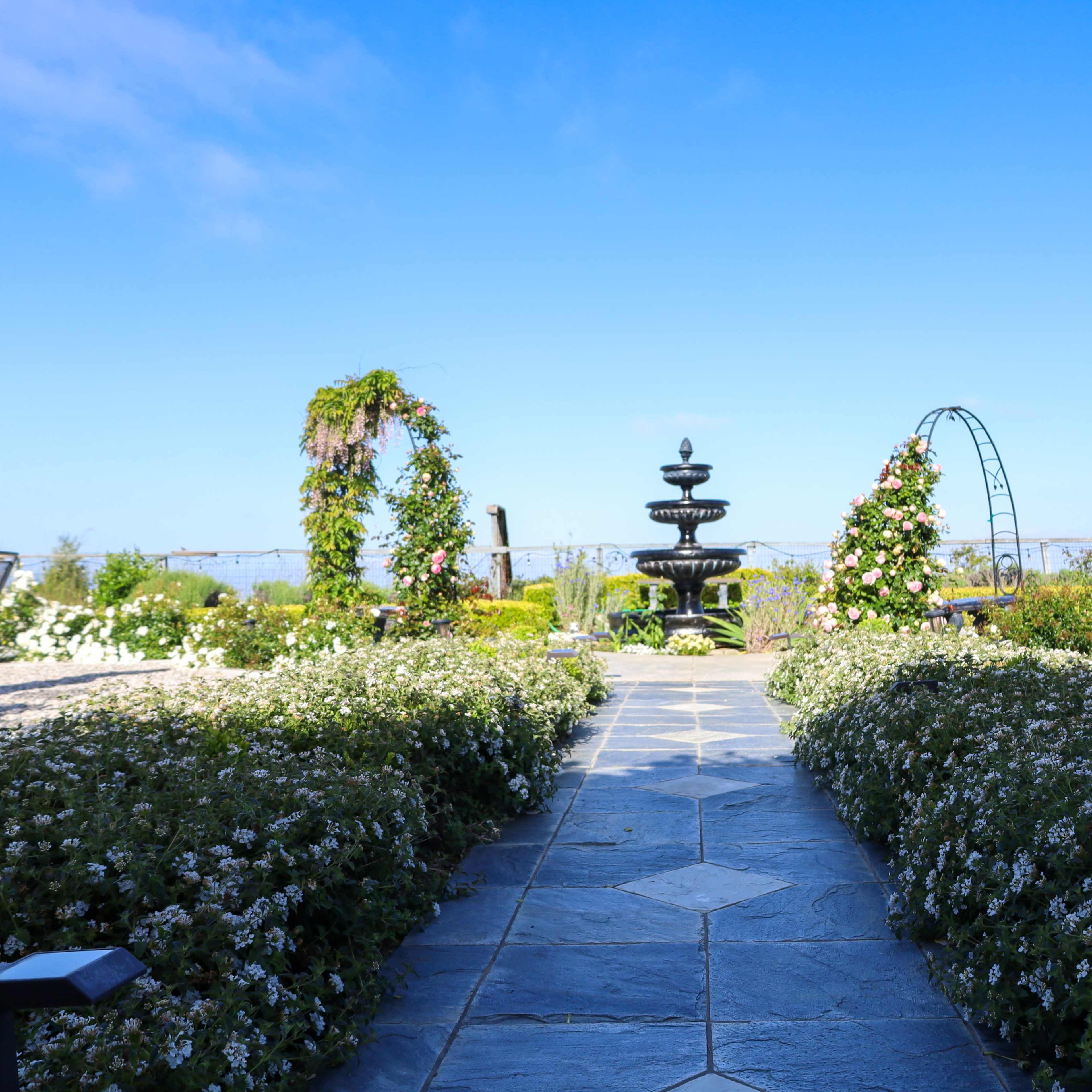 A stone pathway leads through flowerbeds to a central fountain surrounded by arches adorned with greenery under a clear blue sky.