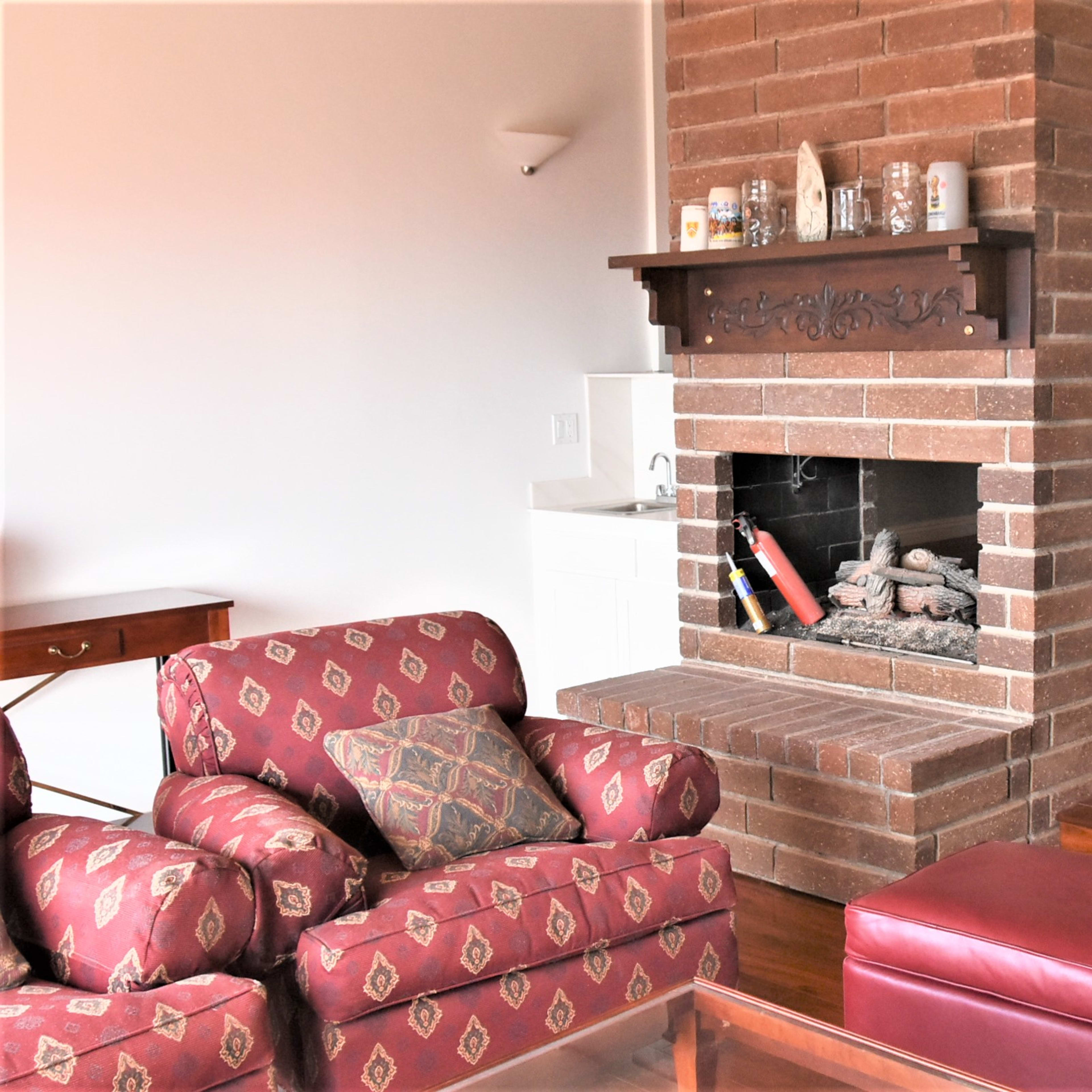 A living room with a brick fireplace surrounded by a red patterned sofa and a wooden dining area in the background.