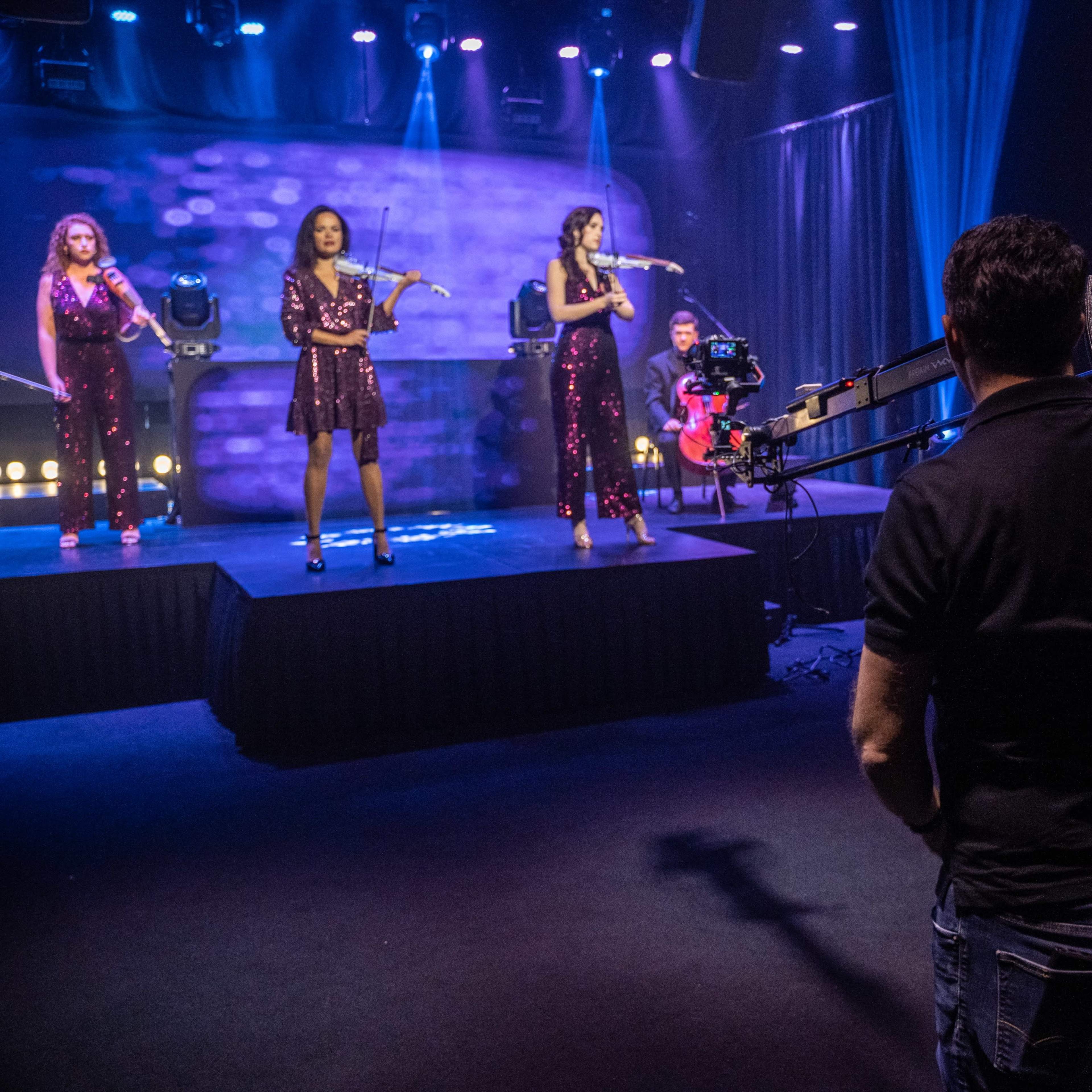 A production crew operates a camera as three performers sing on stage with colorful lighting in a studio setting.