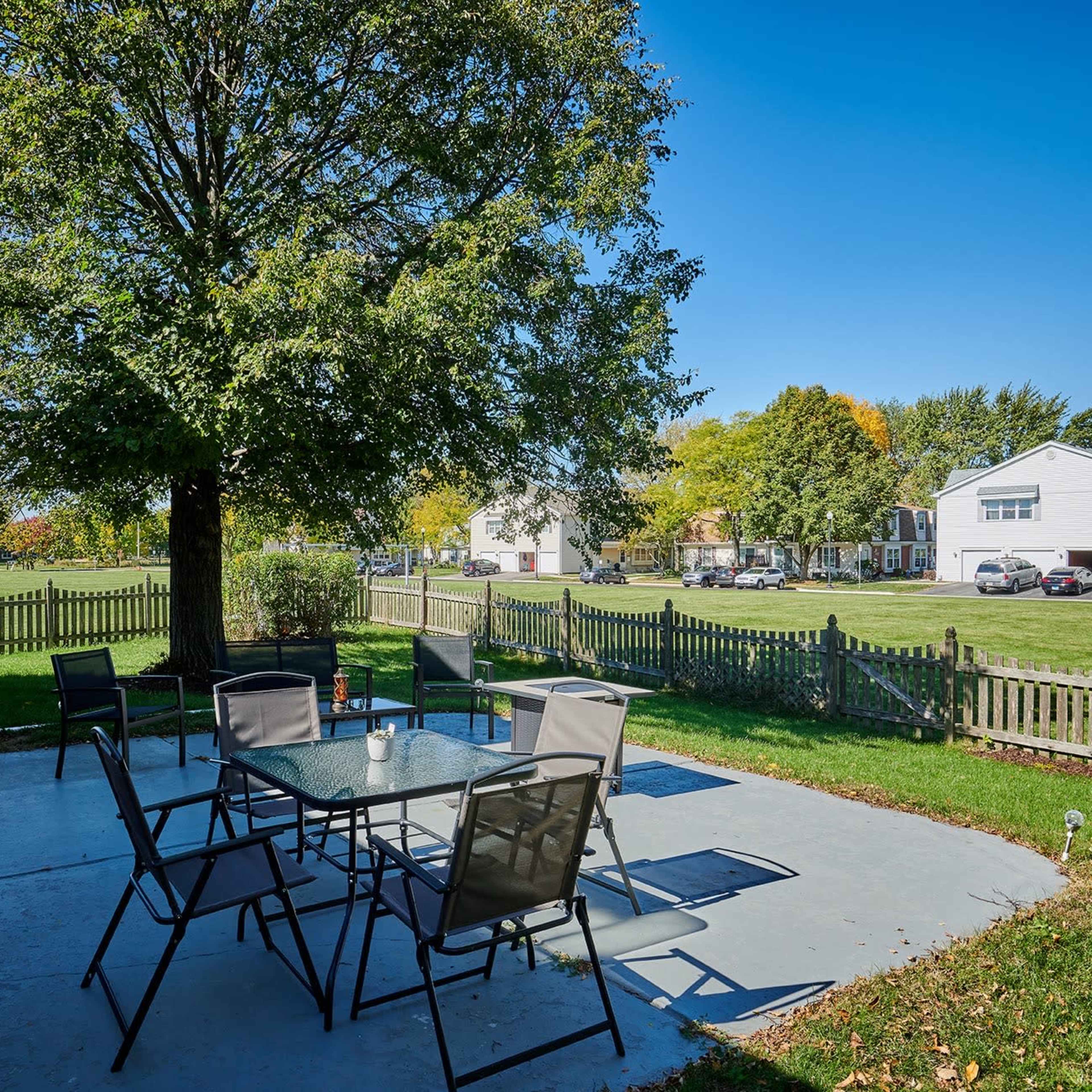 A patio area features a glass table surrounded by chairs, with a large tree nearby and a view of houses and a grassy field in the background.