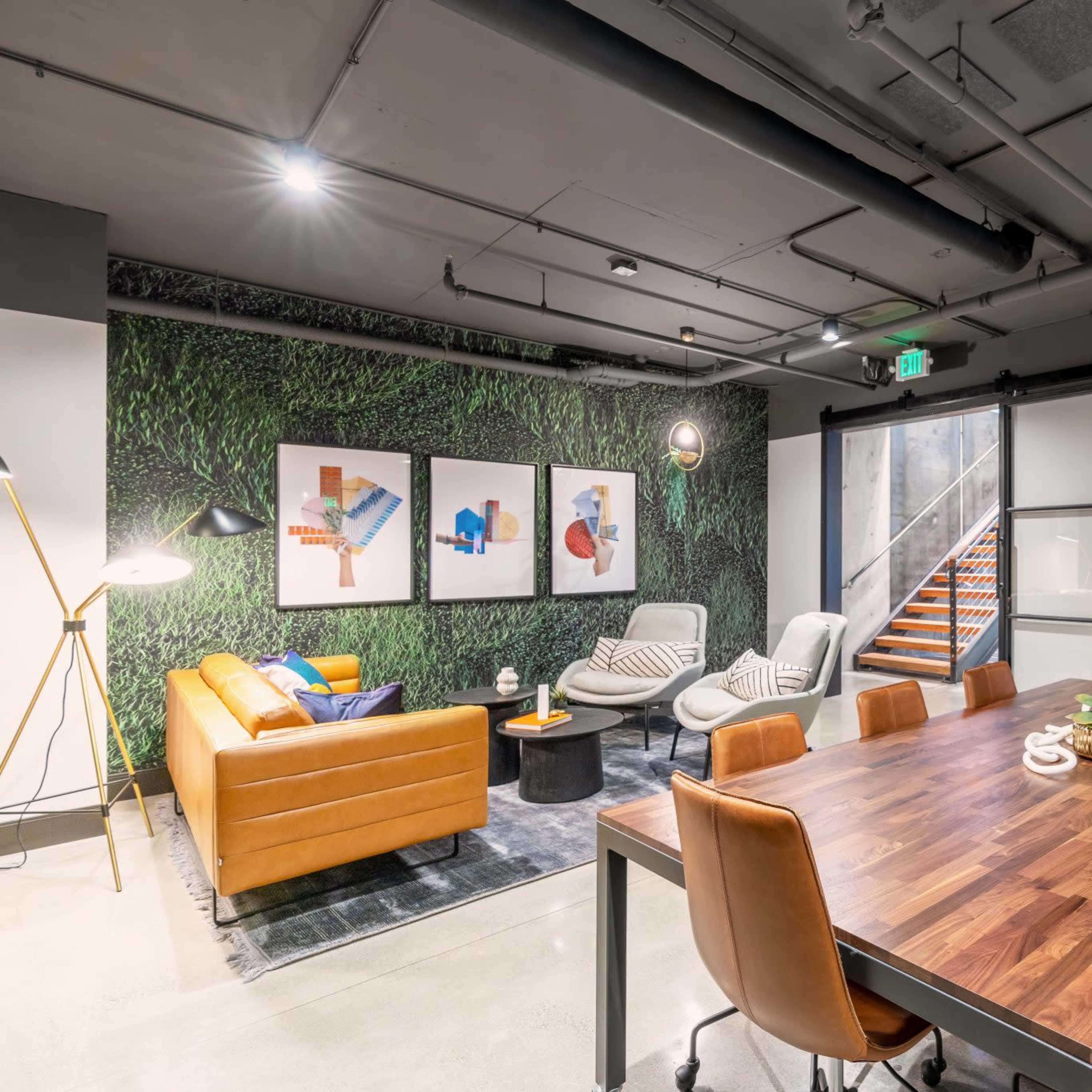 A modern office space featuring a wooden dining table with brown chairs, a lounge area with a leather sofa, and a green textured wall with framed artwork.
