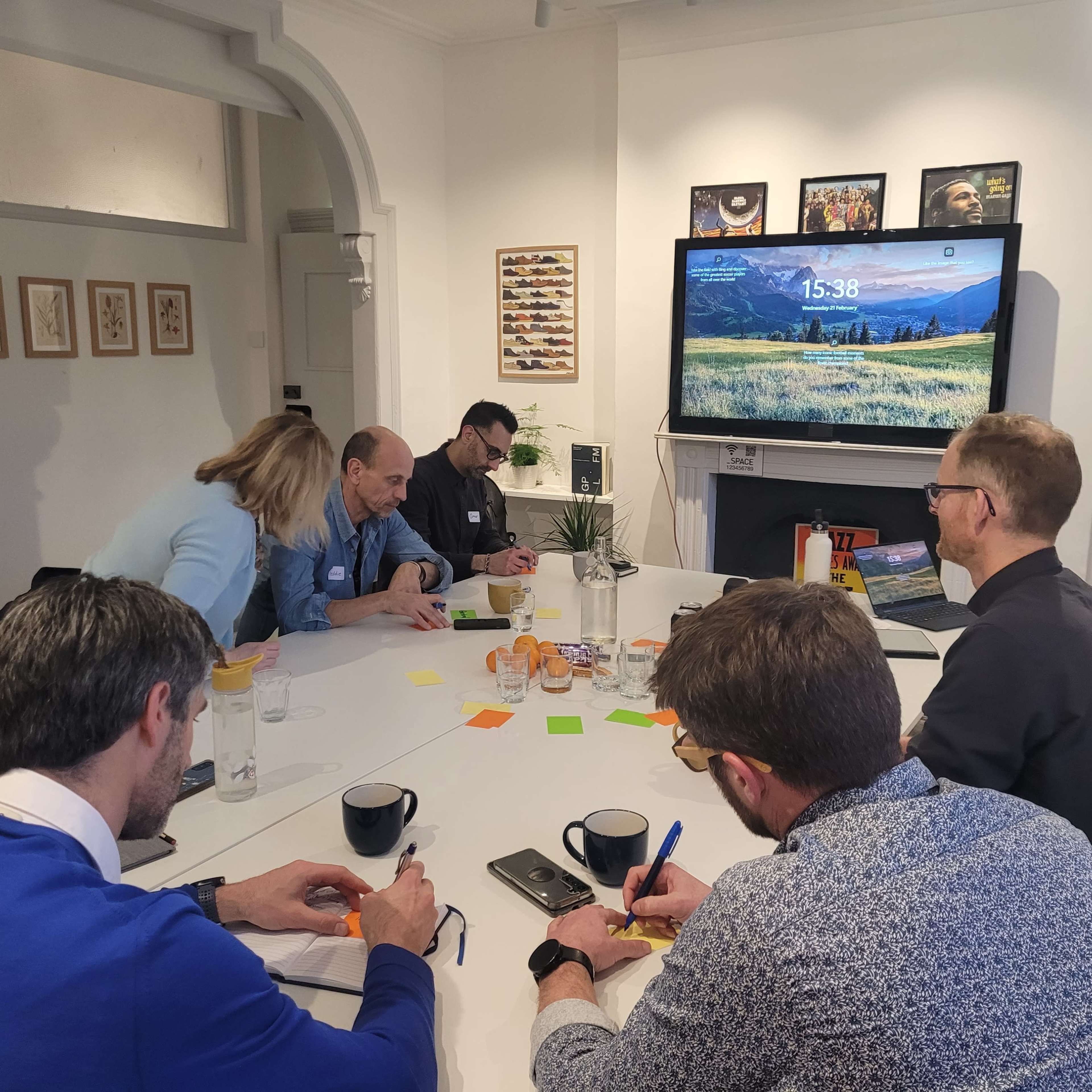 A group of people sit around a table in a meeting room, engaged in discussion and taking notes, with a large screen displaying the time and visuals of a landscape.