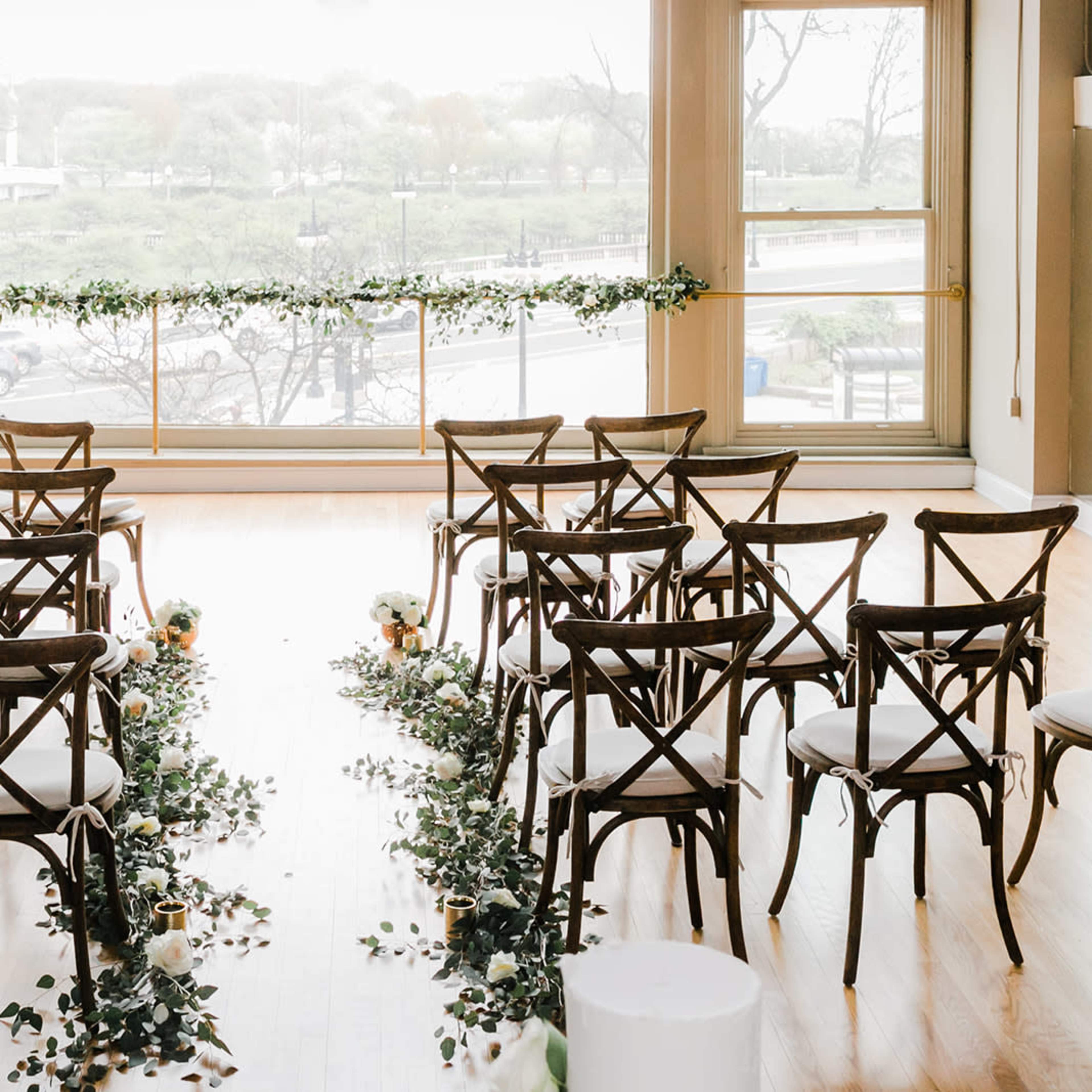 A wedding ceremony setup featuring rows of wooden chairs flanking a floral aisle in a bright room with large windows overlooking a green landscape.