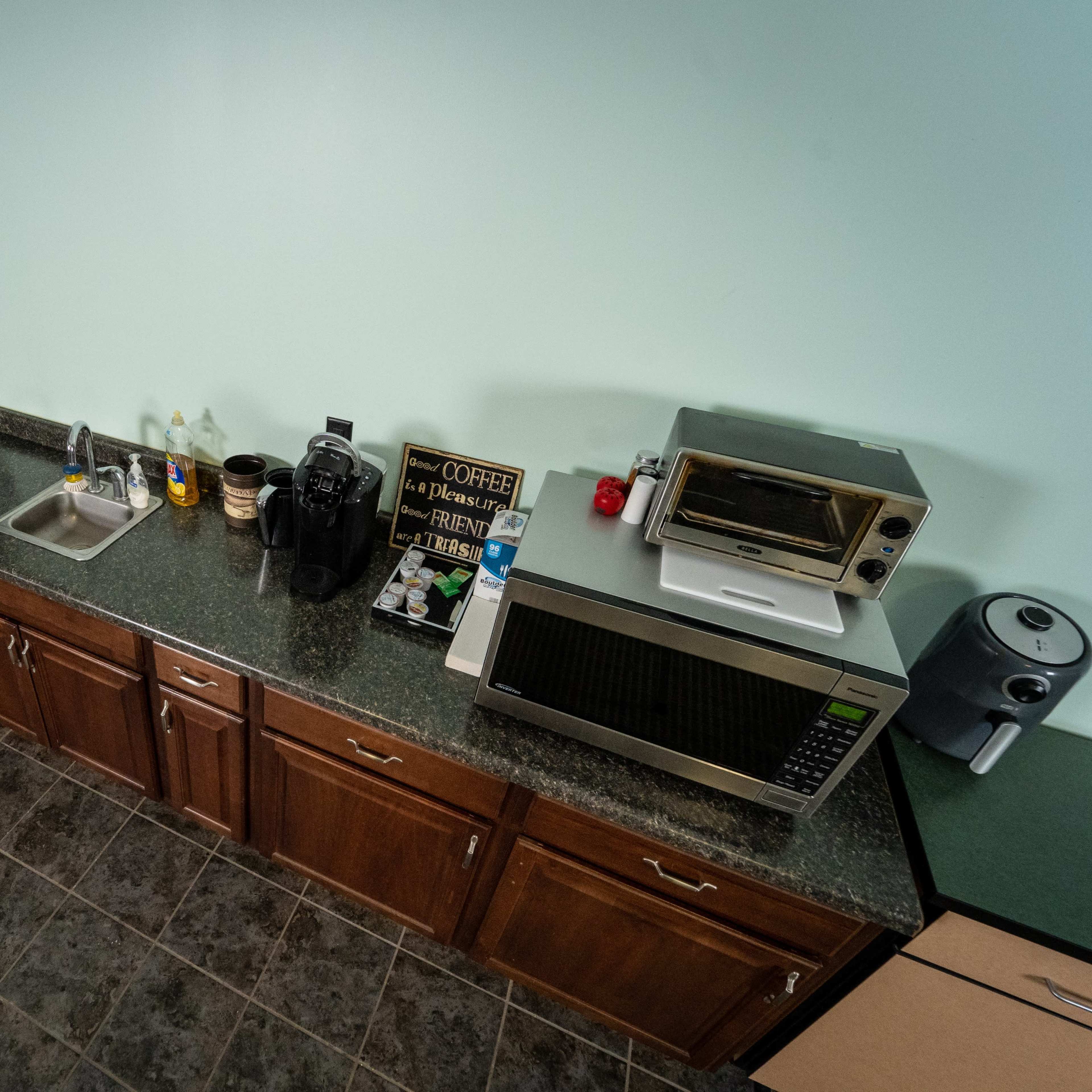 A kitchen countertop with a sink, coffee maker, toaster oven, microwave, and various utensils, set against a light blue wall and a dark tile floor.