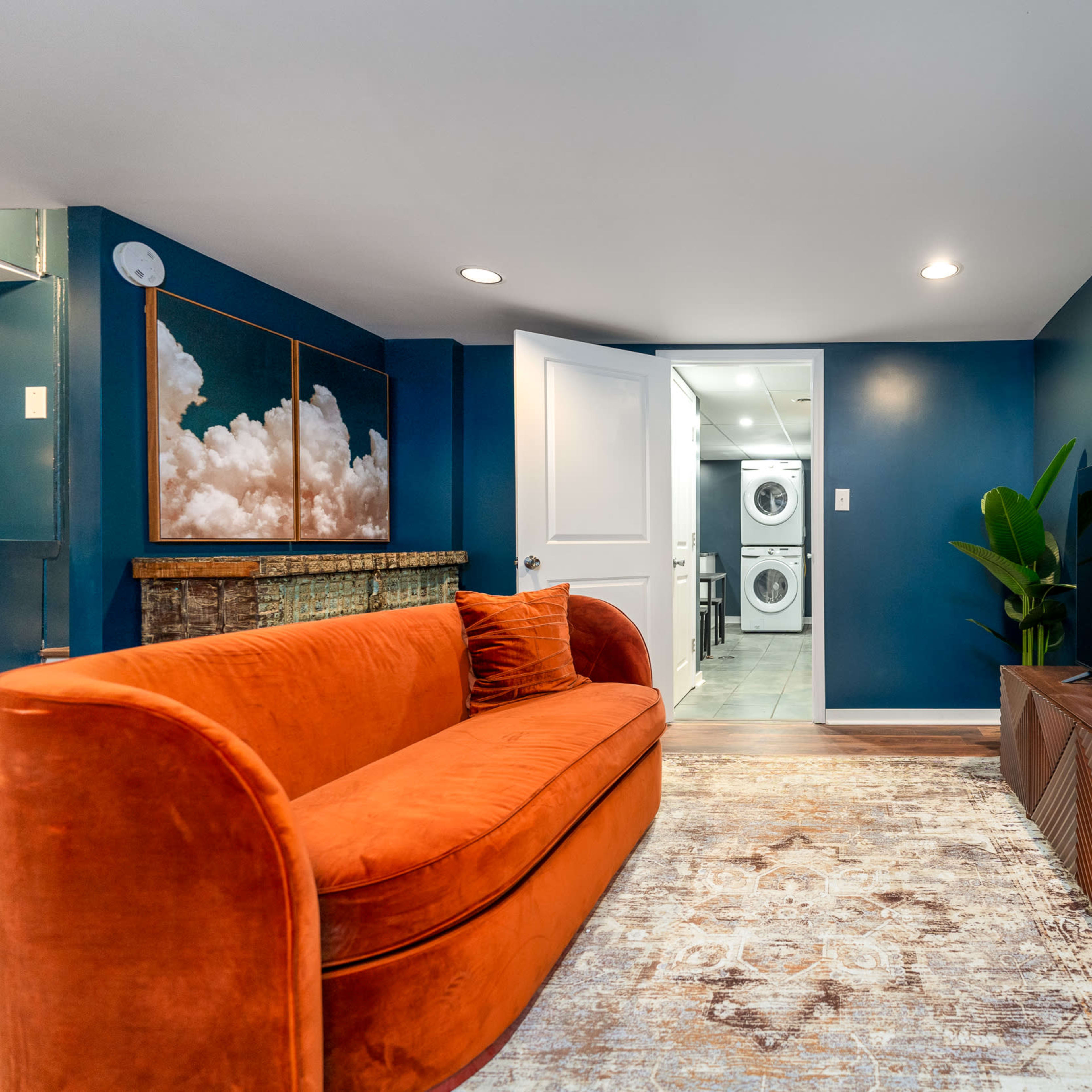A bright blue room features an orange sofa, a wooden TV stand, and a doorway leading to a laundry area.