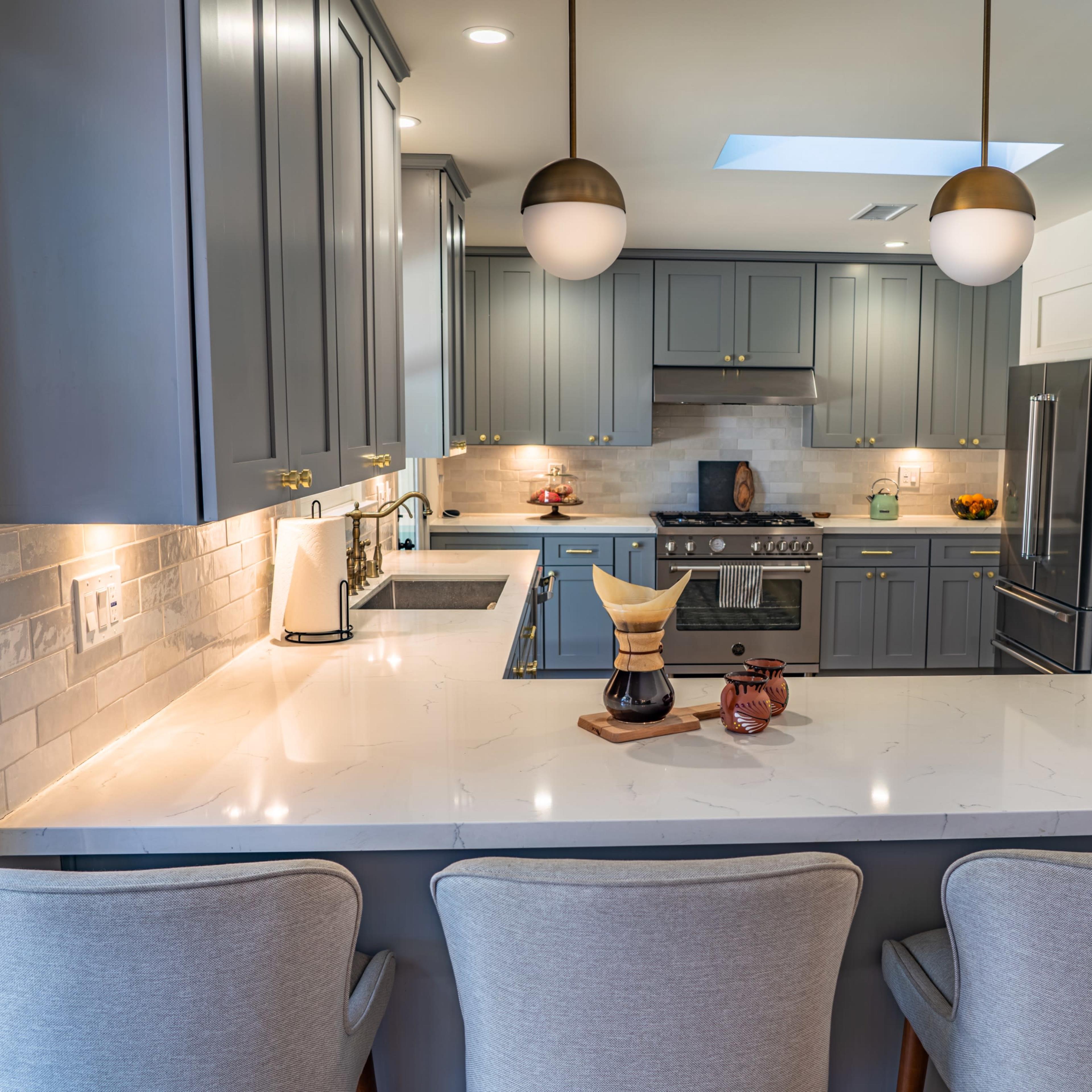 A modern kitchen features gray cabinets, a white countertop, and stainless steel appliances, with three bar stools positioned at a central island.