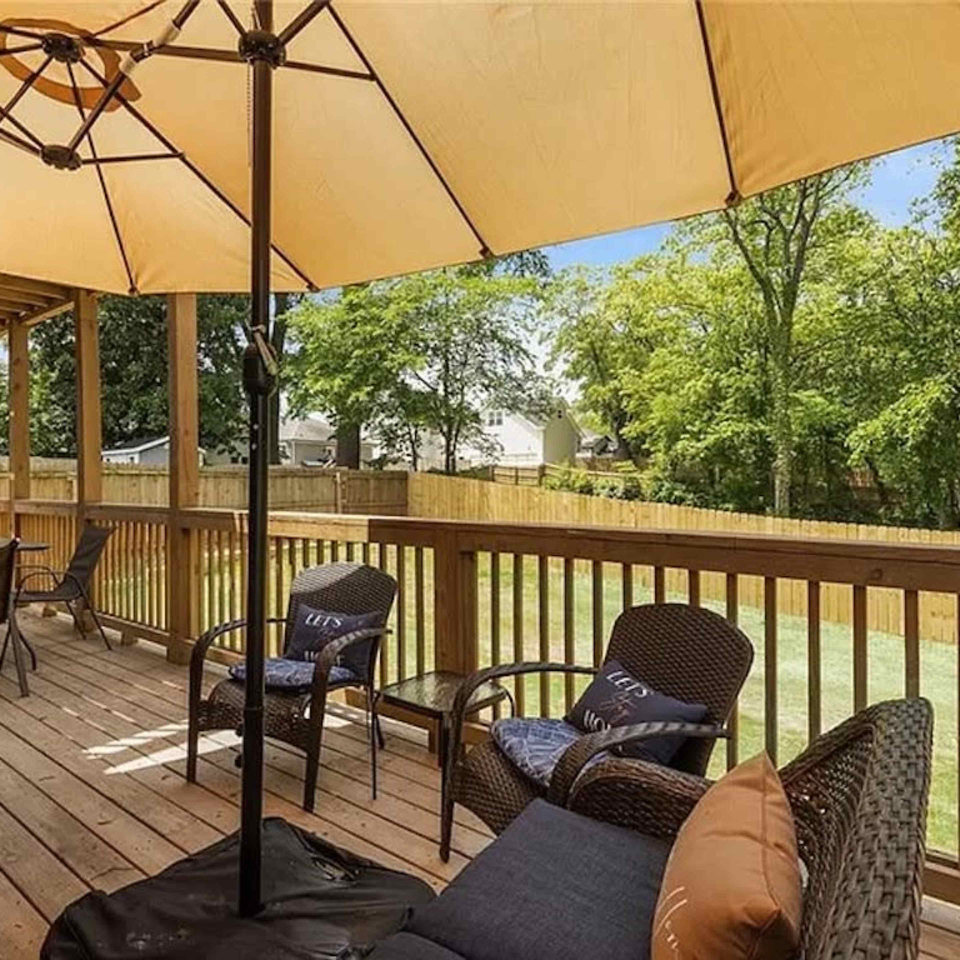 A wooden deck with an umbrella and outdoor seating, overlooking a grassy backyard and trees.
