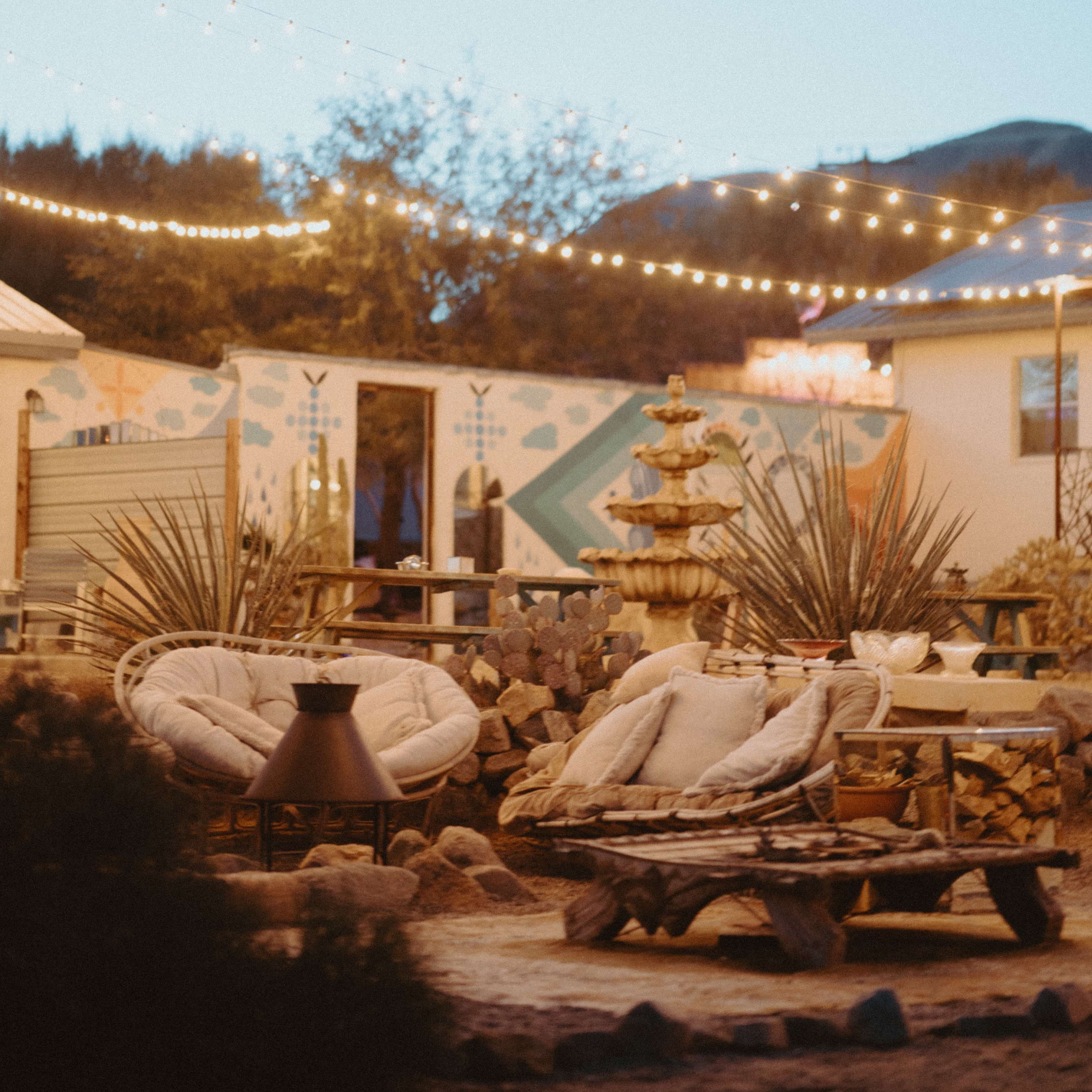 A cozy outdoor seating area with two circular chairs, a small fire pit, and string lights illuminating the surrounding desert landscape.