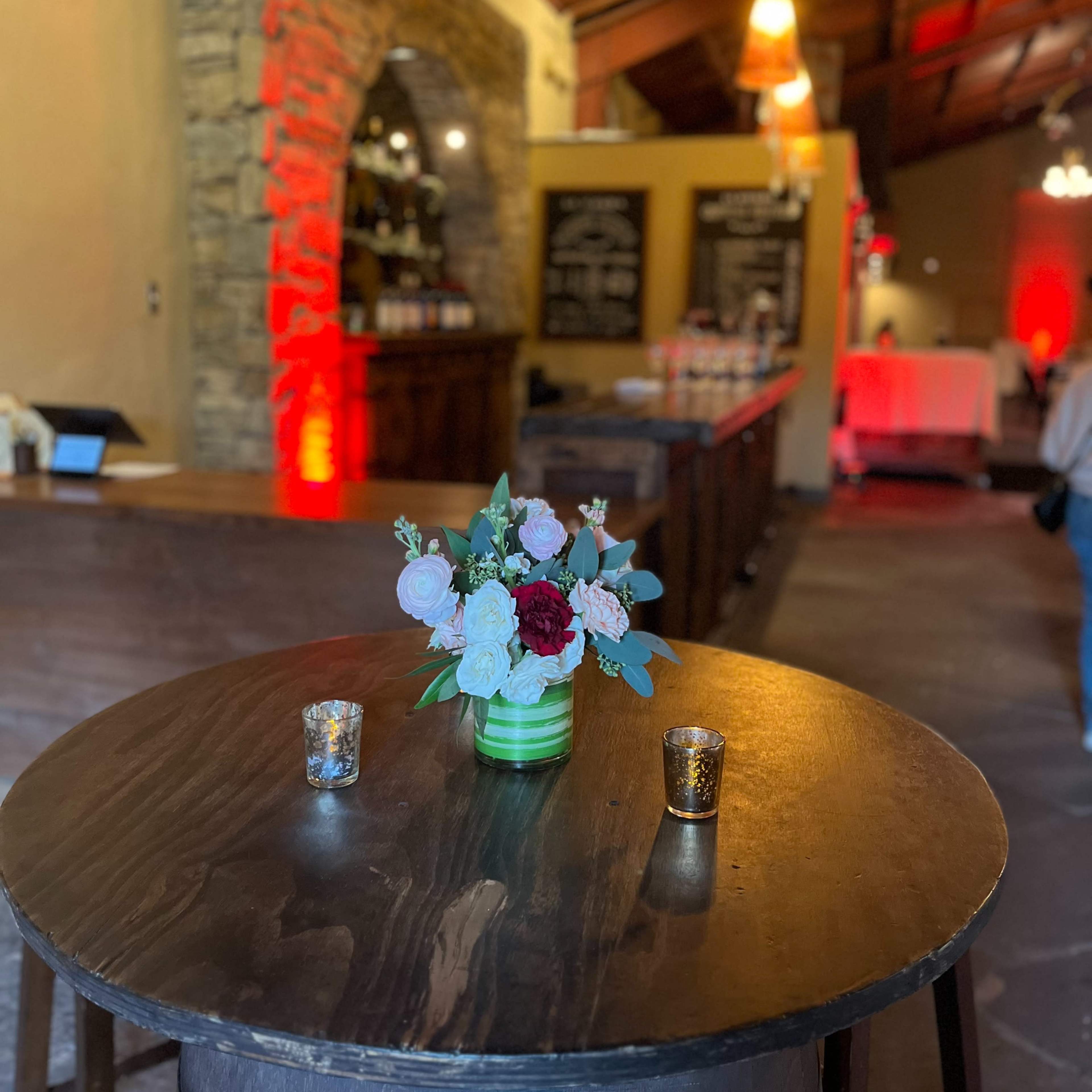 A small round table with a floral arrangement and candles is set in front of a bar area in a rustic venue.