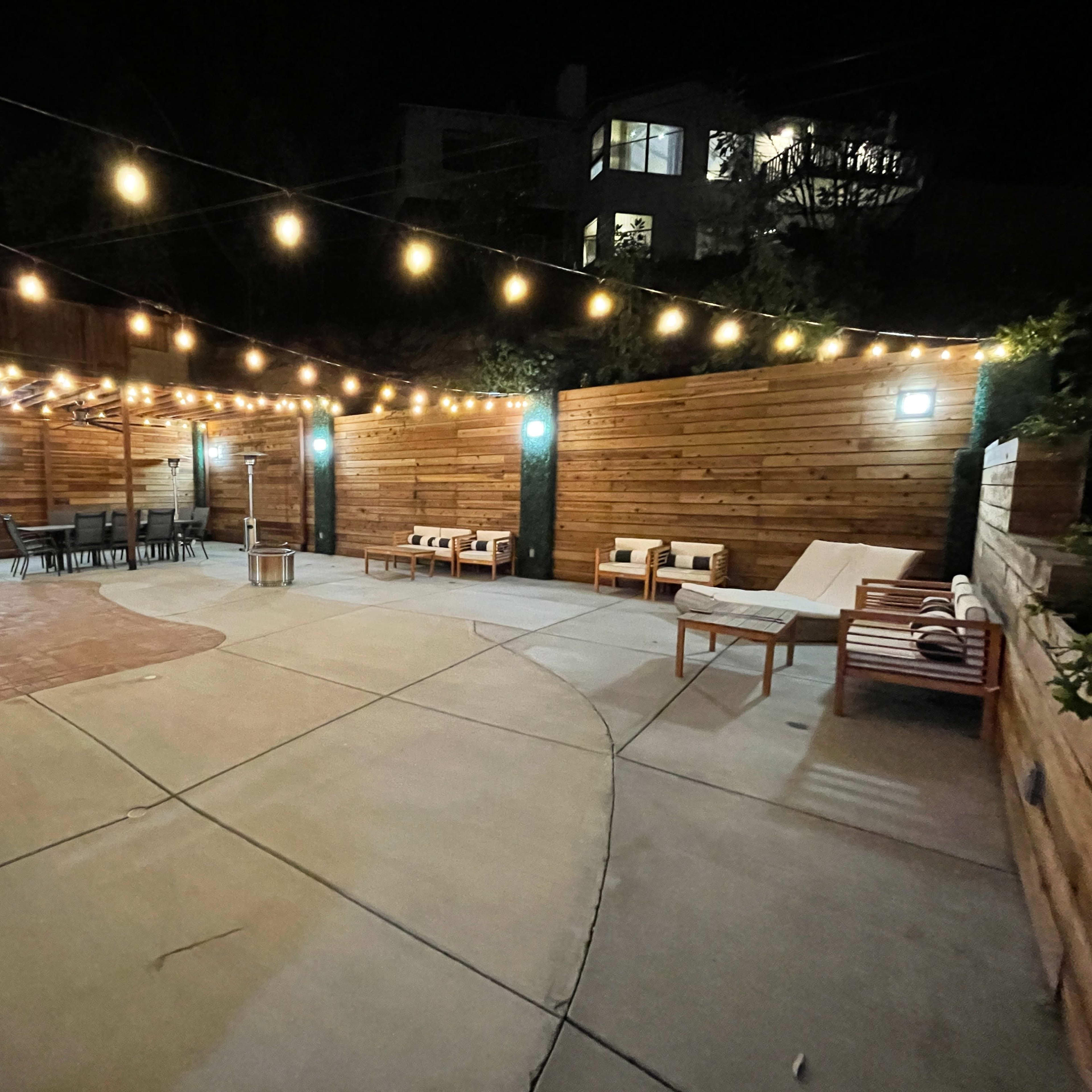 A night scene of a spacious outdoor patio area with string lights above, featuring wooden furniture and a dining table.