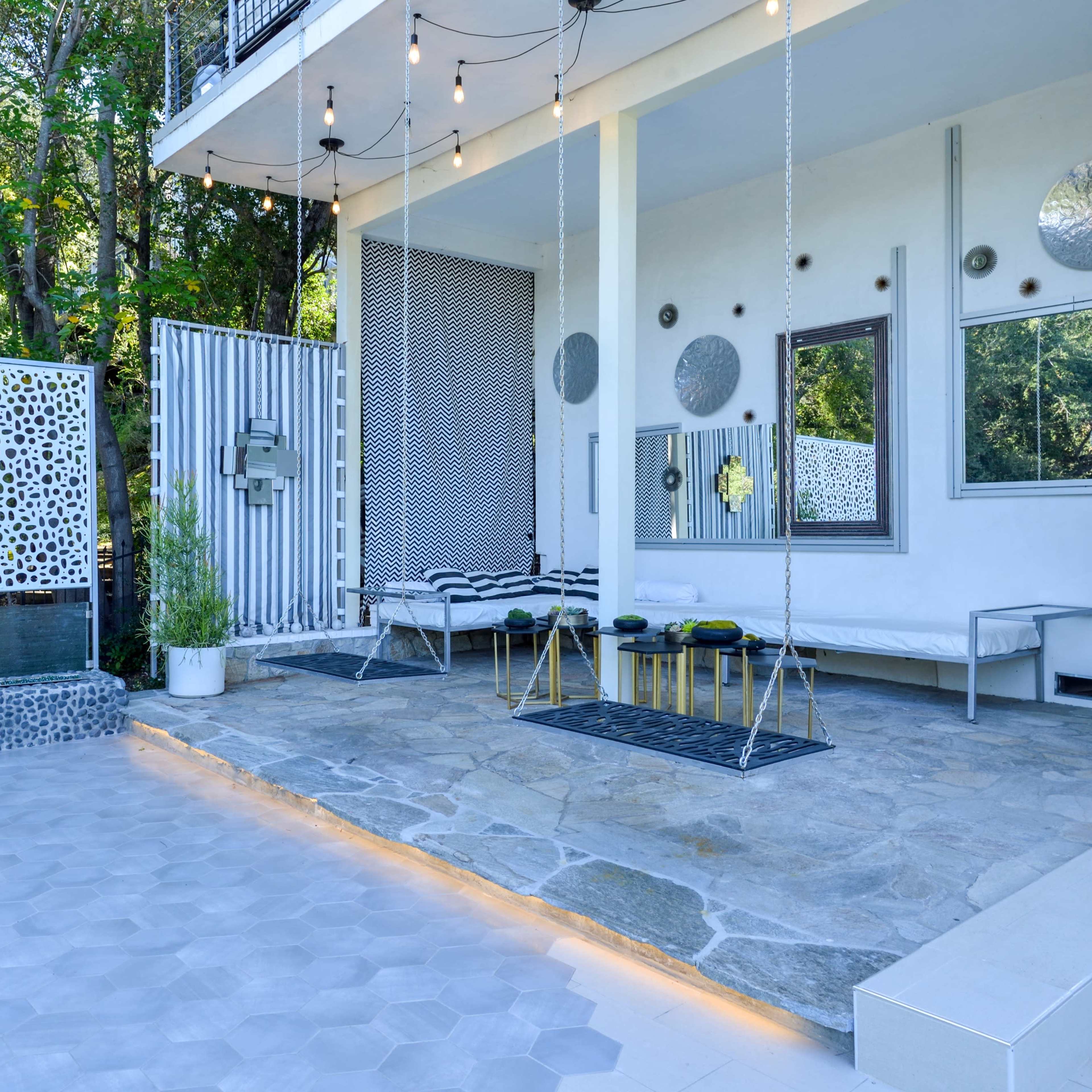 A modern outdoor patio area featuring a stone floor, decorative wall screens, hanging lights, and various seating arrangements amid greenery.