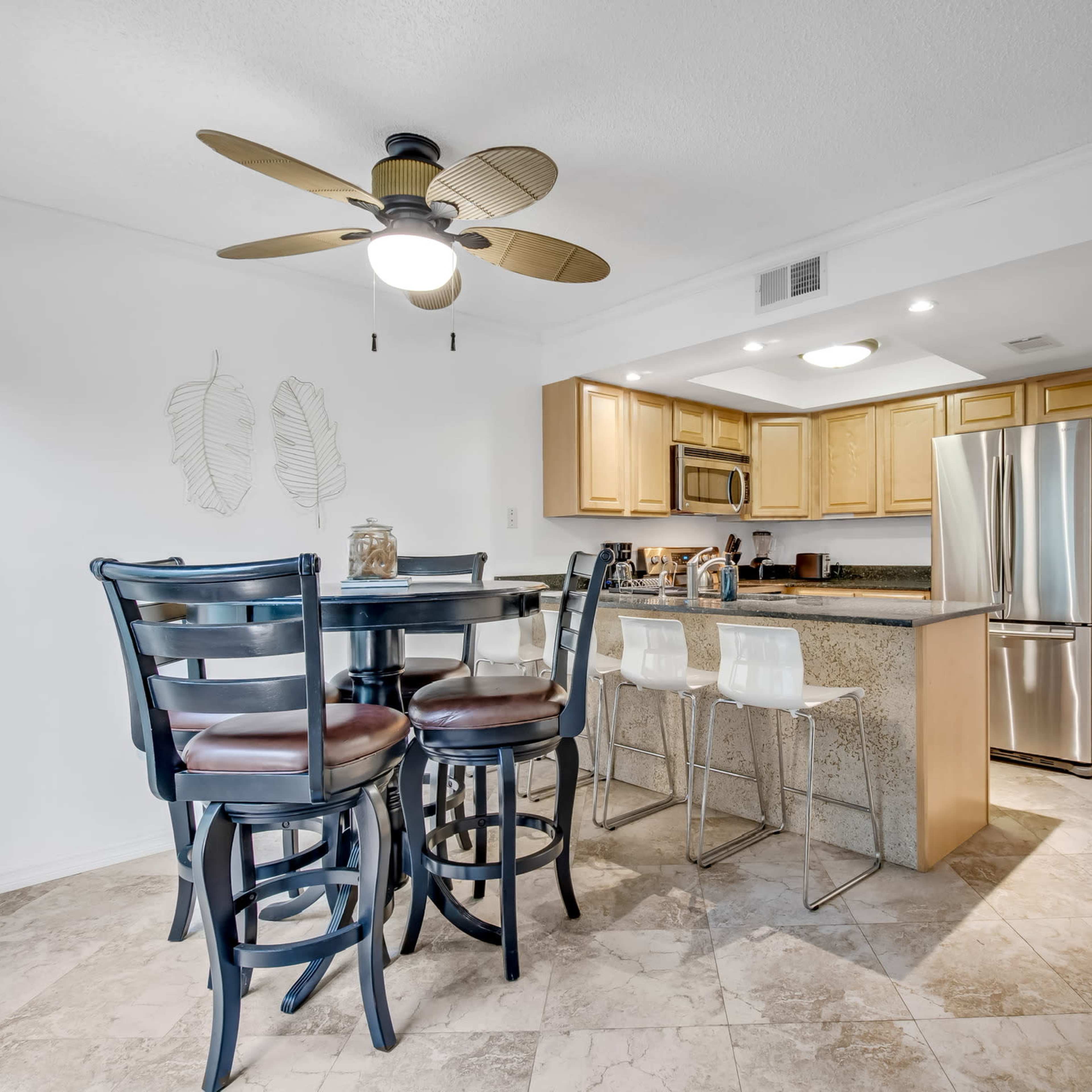 A kitchen-dining area features a round wooden table with black chairs, a bar with clear stools, and wooden cabinets against a granite countertop.