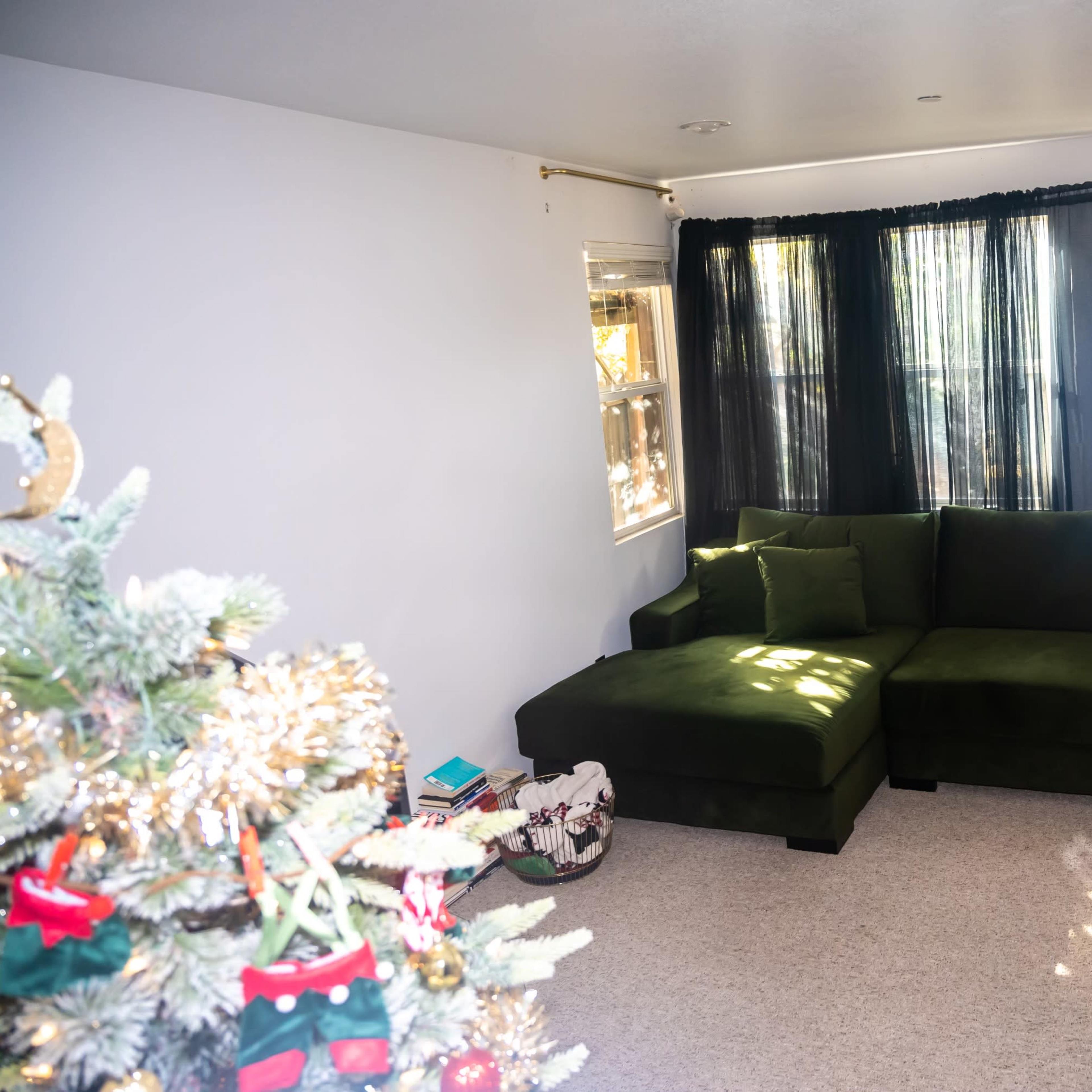 A green sectional sofa positioned in a brightly lit living room with large windows and a Christmas tree in the foreground.