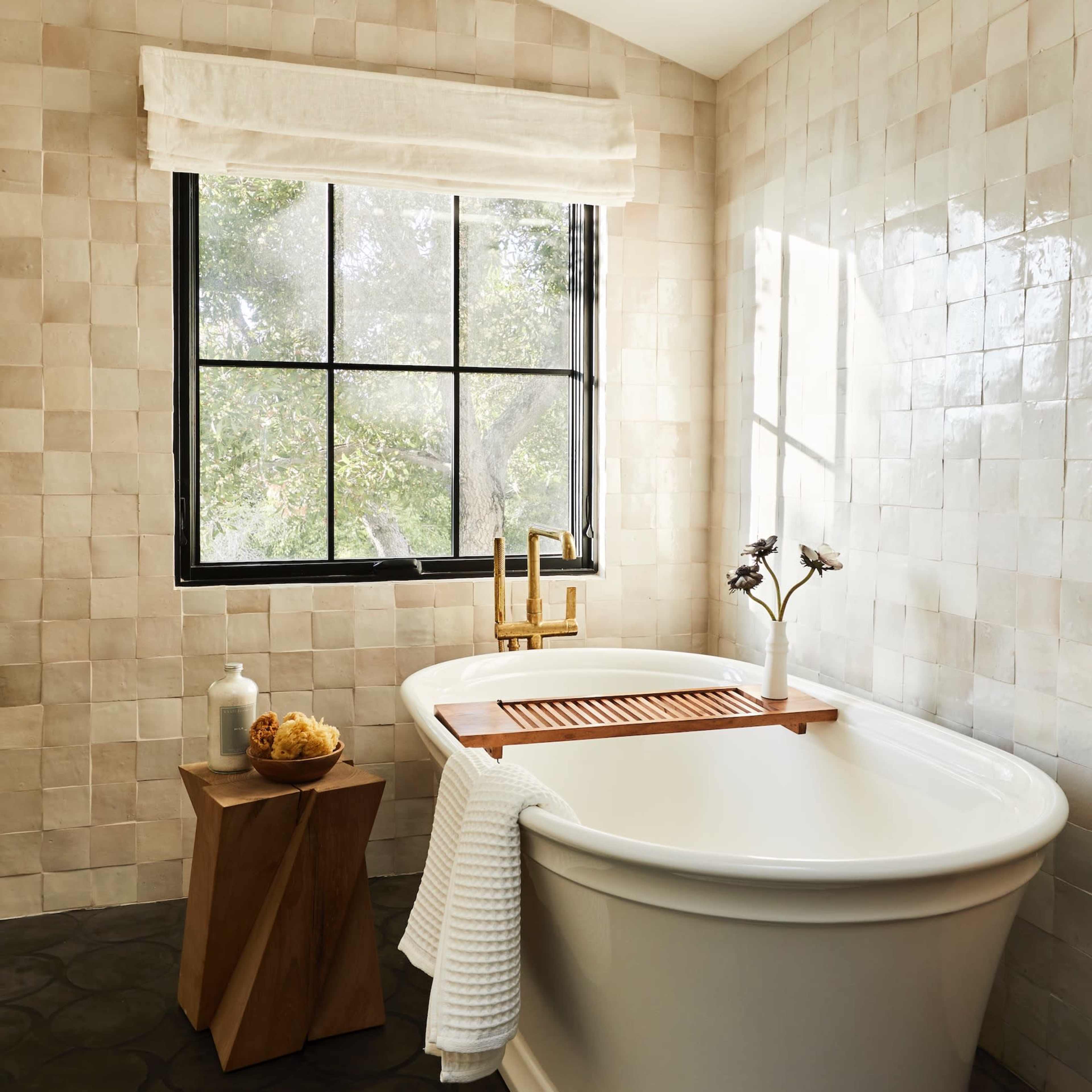 A white freestanding bathtub is positioned next to a window, accompanied by a wooden side table and a towel.