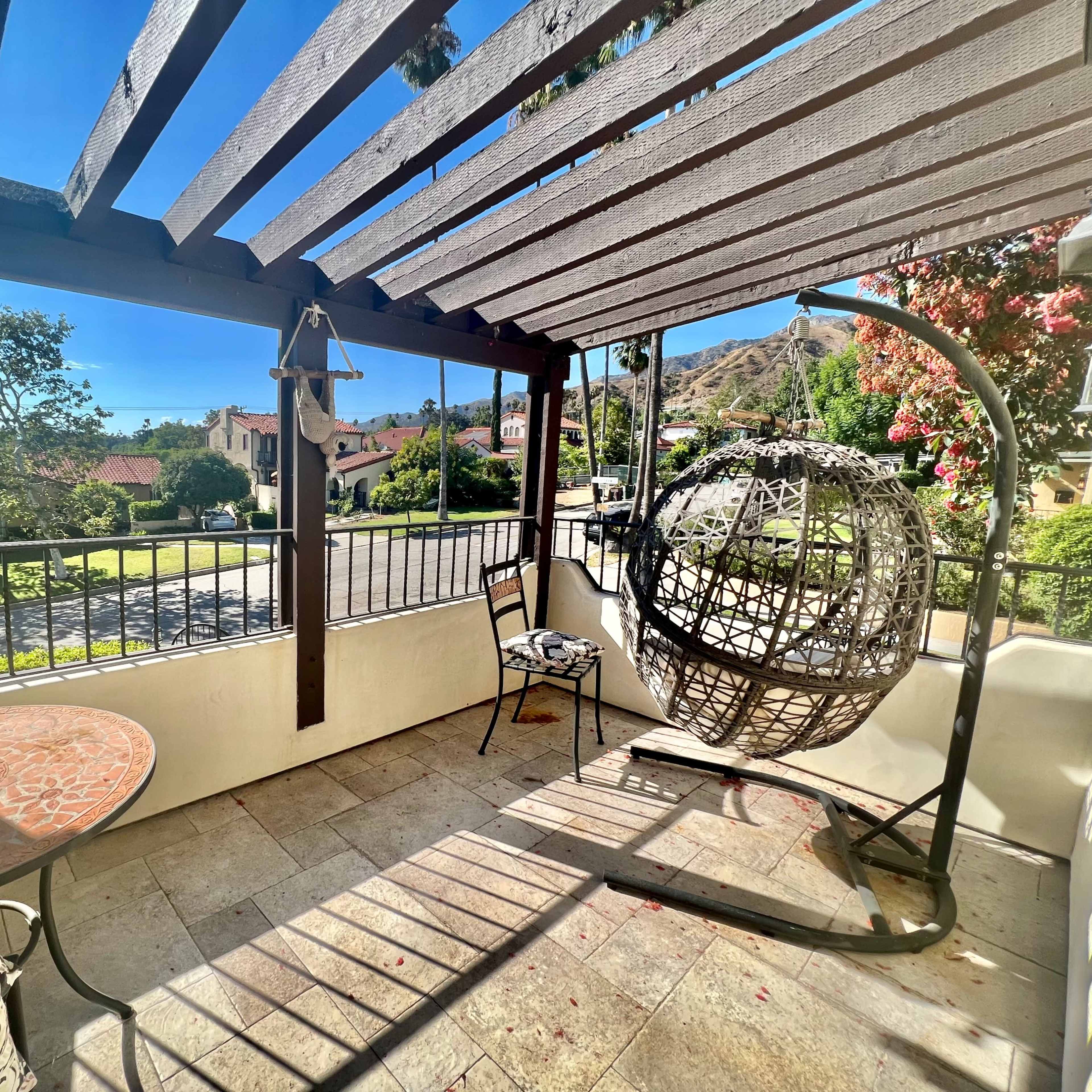 A covered patio features a circular hanging chair and a small table, with a view of greenery and houses beyond.
