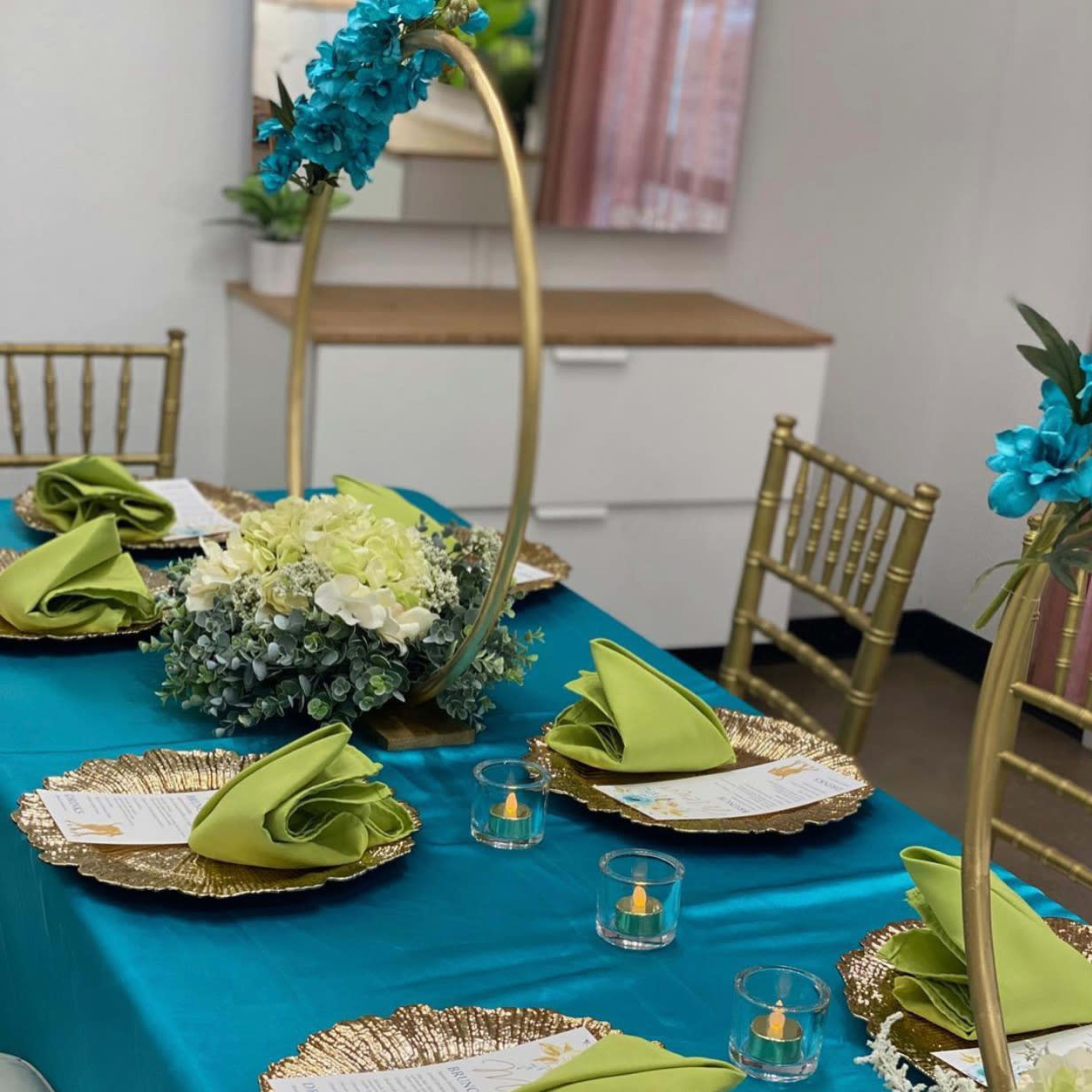 A neatly arranged dining table features gold chargers, green napkins, and a floral centerpiece, with gold chiavari chairs surrounding the setup.