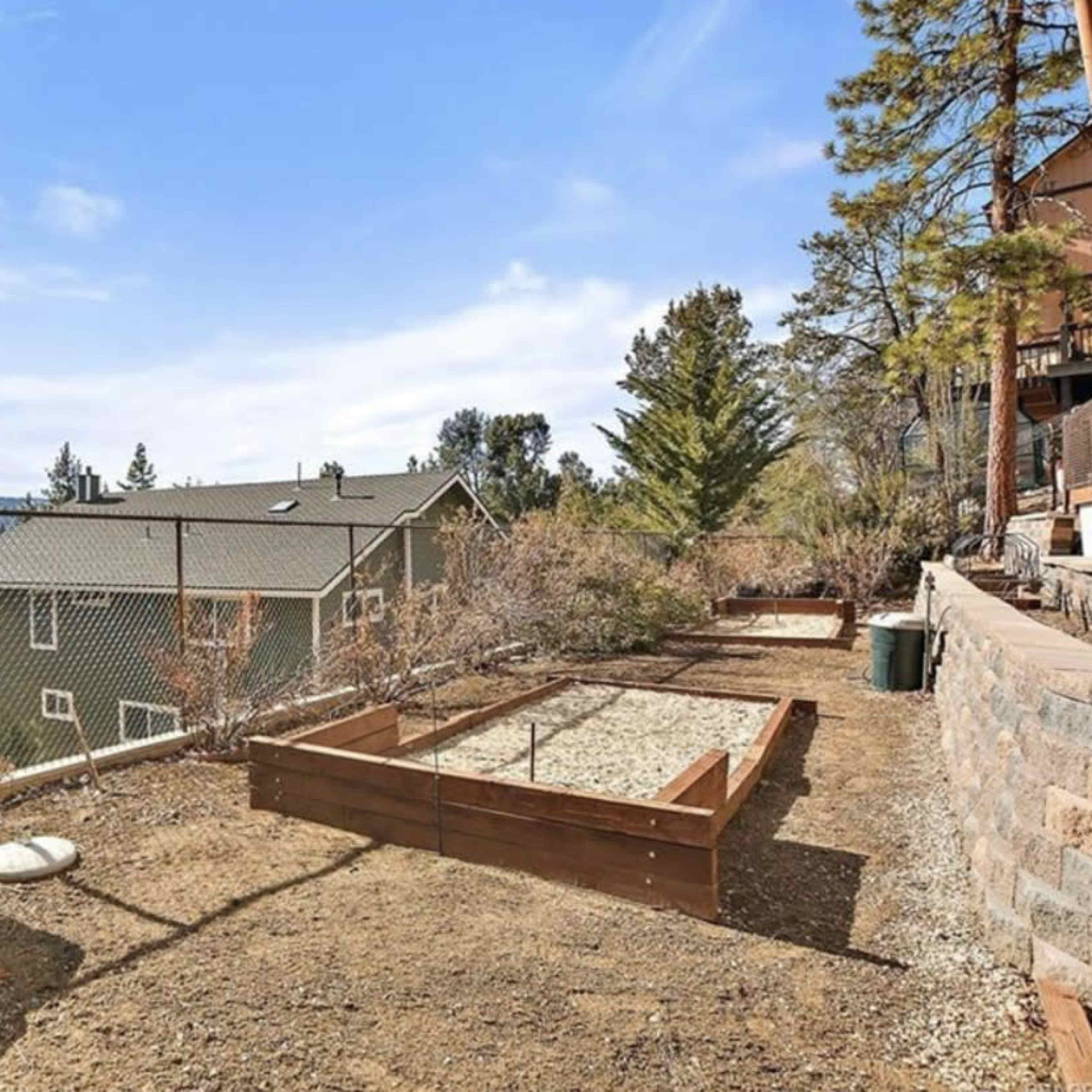A fenced garden area with a raised wooden planter box is set against a backdrop of trees and a neighboring house.