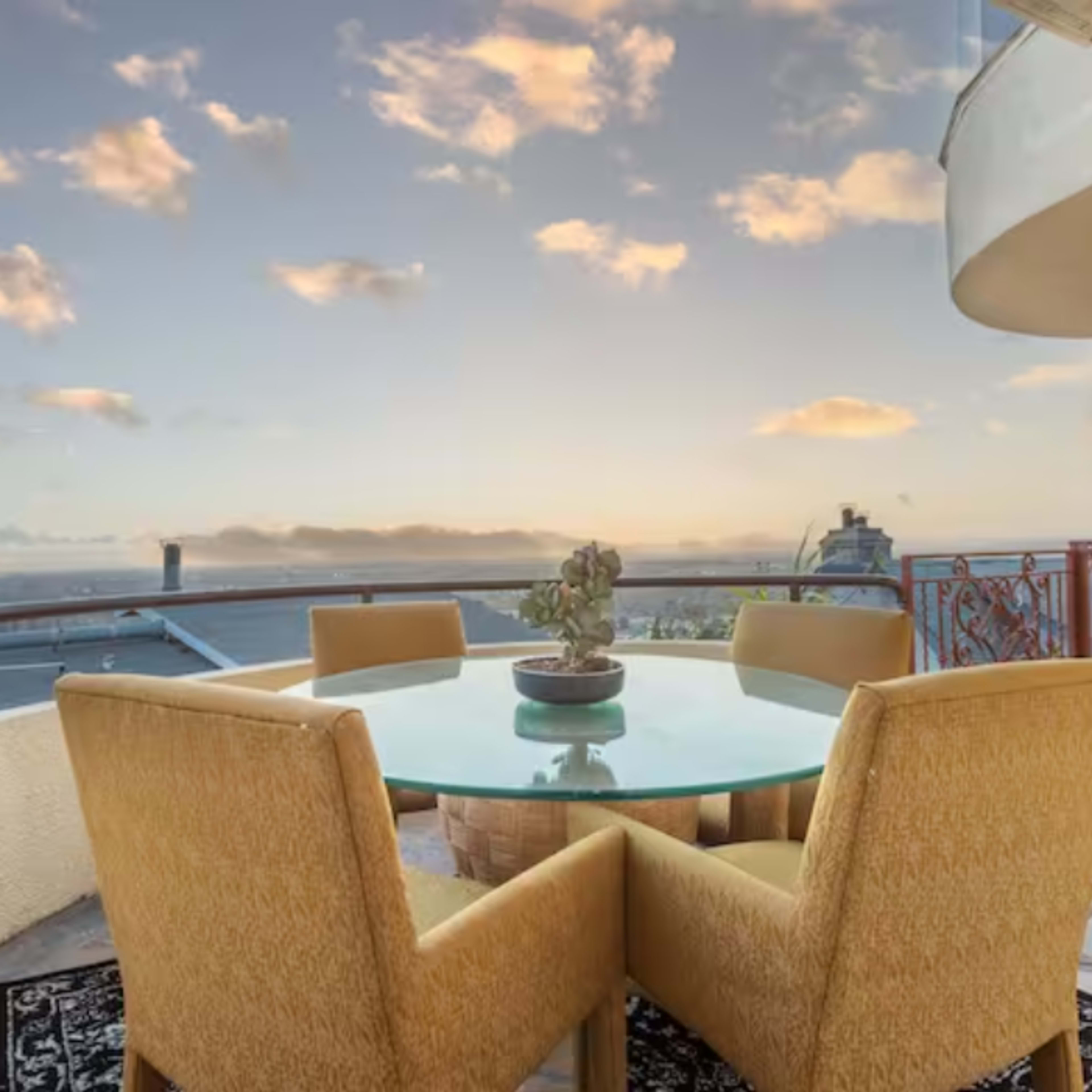 A circular glass table with four yellow chairs is set on a balcony overlooking a sunset sky and distant rooftops.