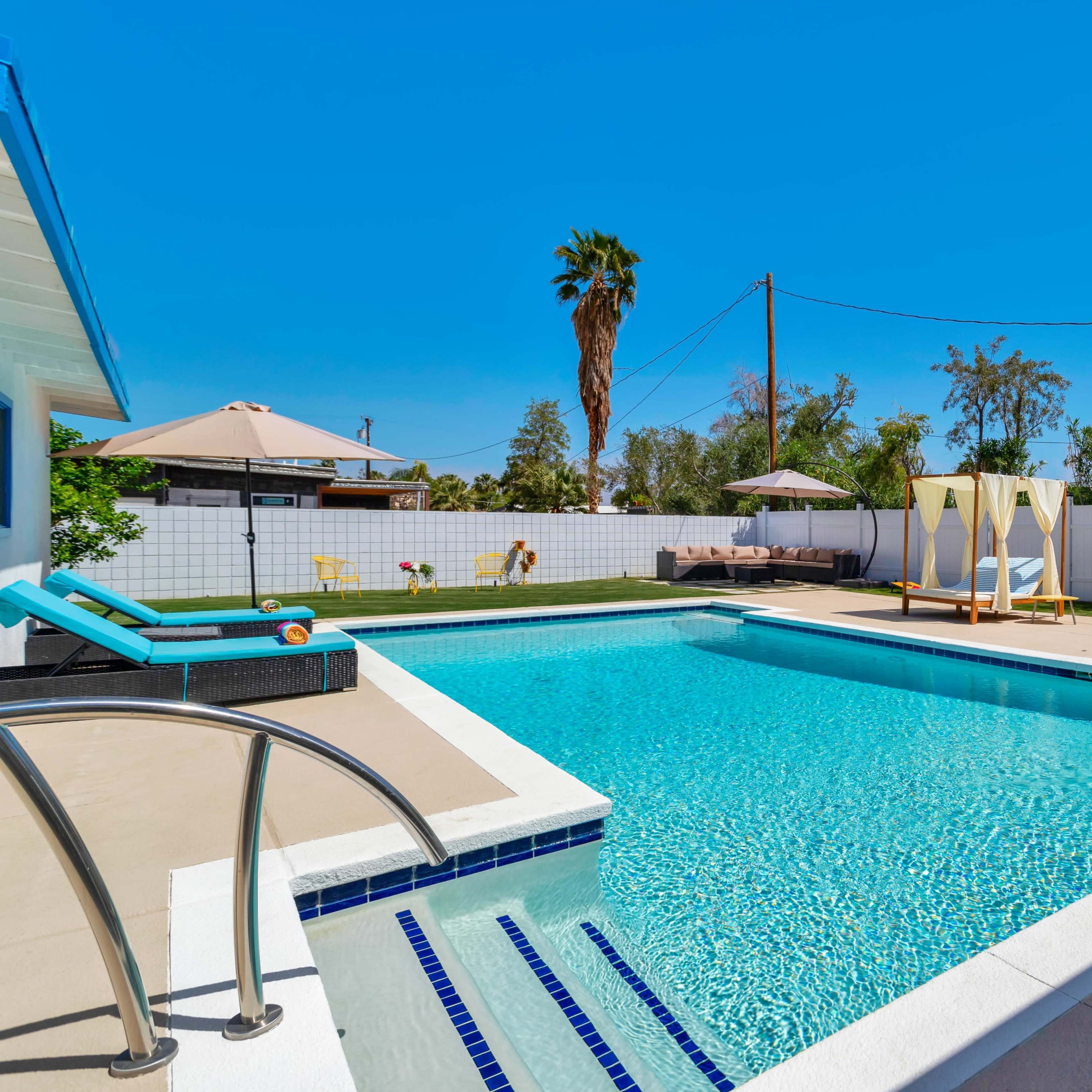 A backyard pool area featuring a large swimming pool, a hot tub, lounge chairs, and a shaded seating area surrounded by palm trees and greenery.