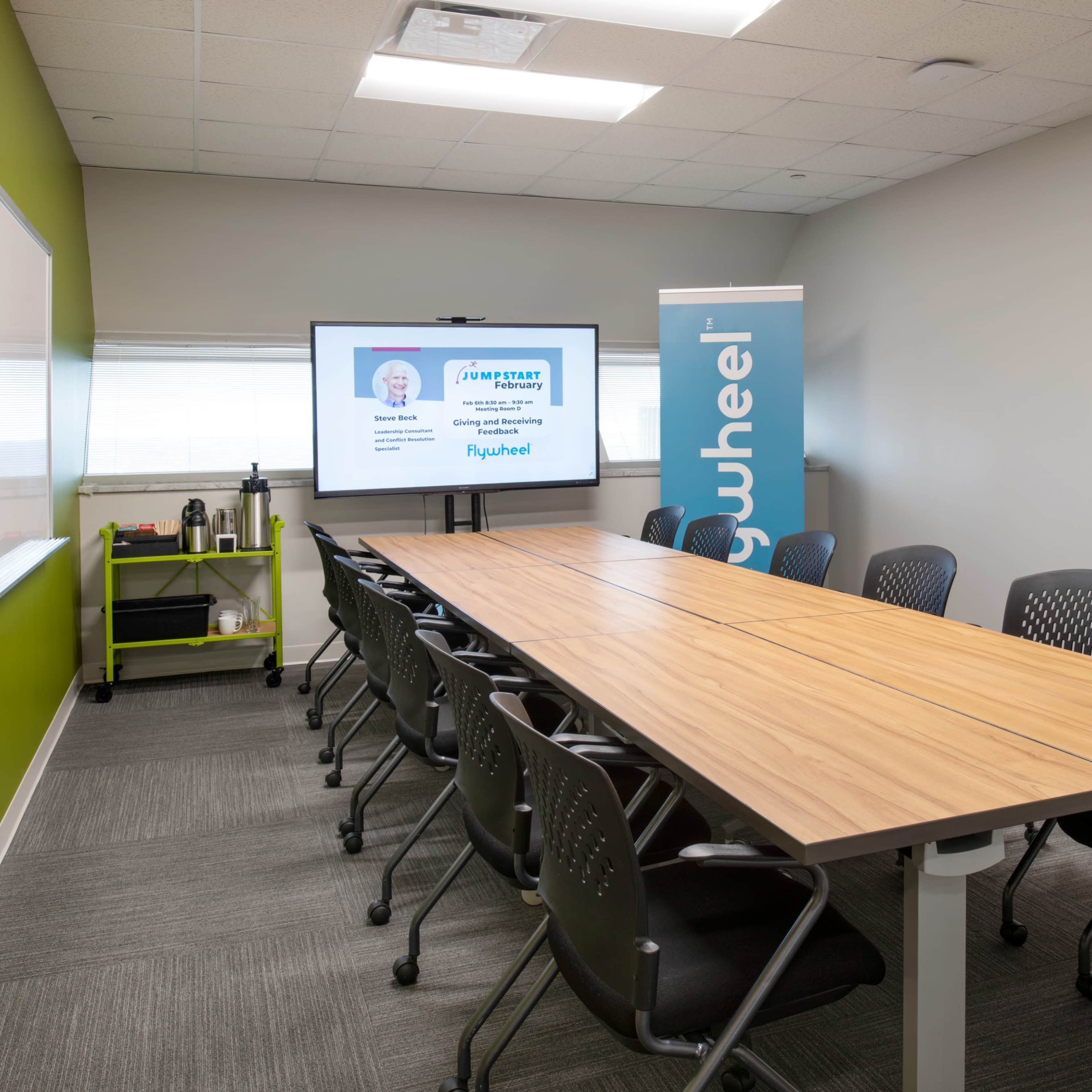 A modern conference room with a long wooden table, office chairs, a whiteboard, and a screen displaying a presentation.