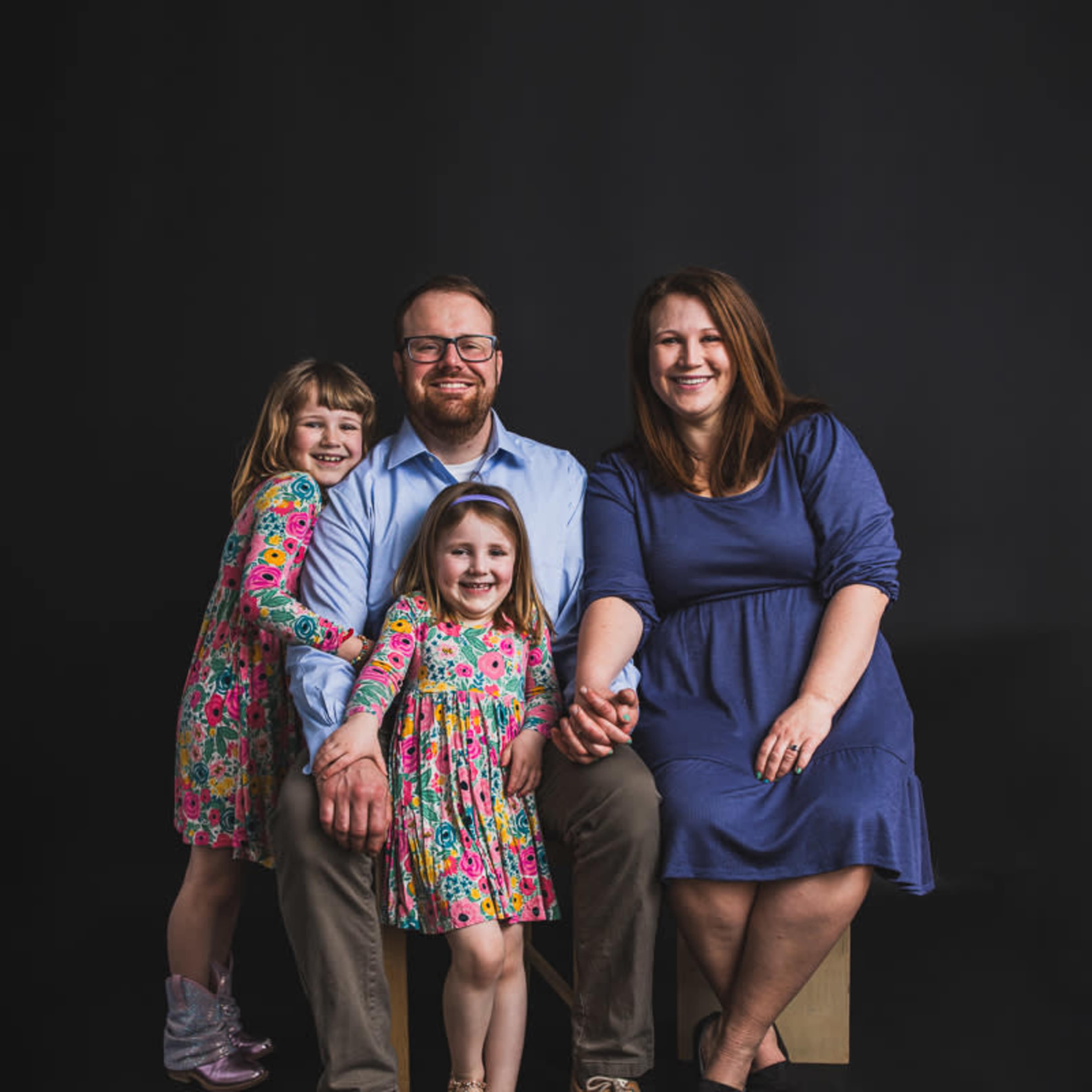 A family of four, including two young girls and their parents, is seated together against a black background, smiling at the camera.