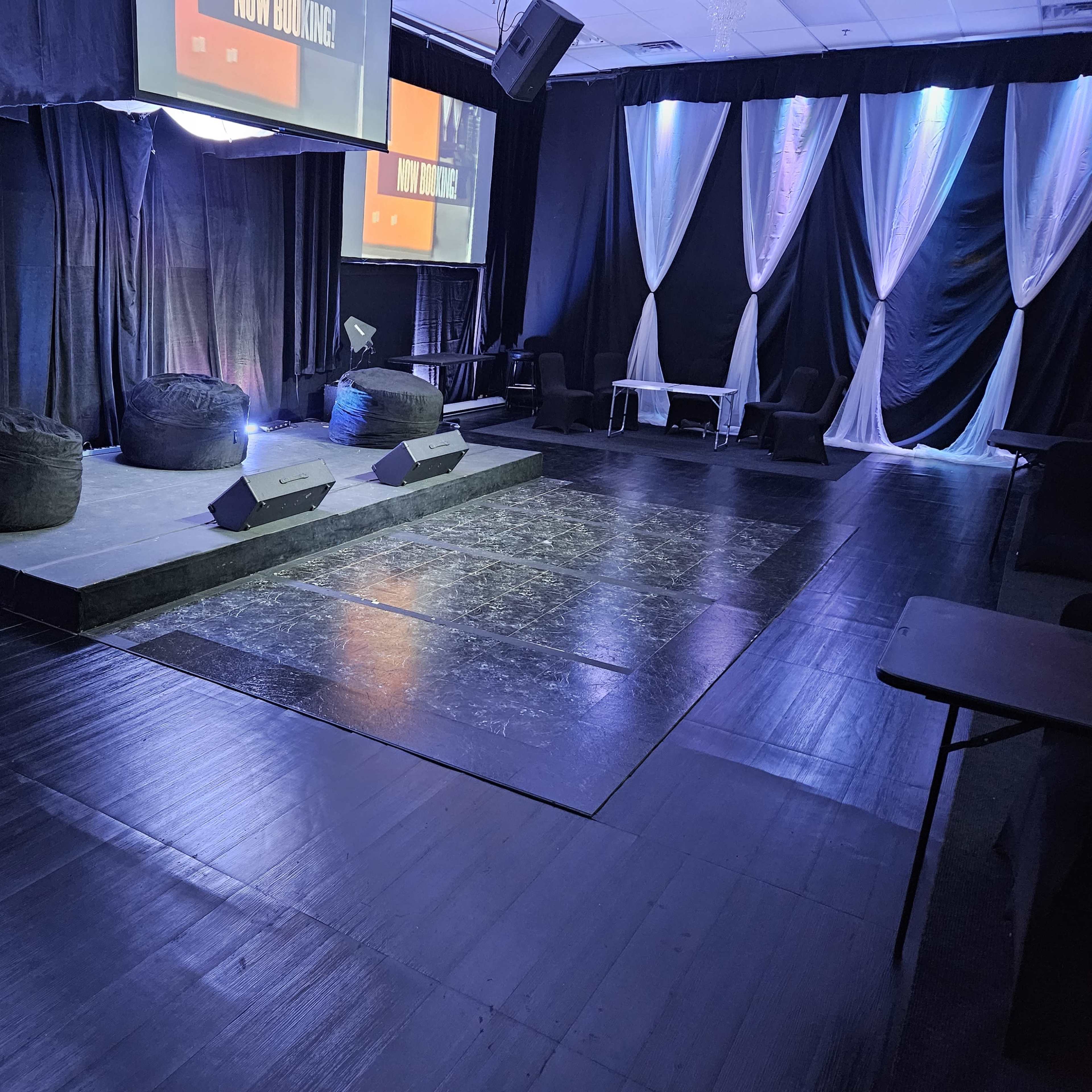 A stage area with three black bean bag chairs, two speaker monitors, and a large screen backdrop, all set within a room draped in black and white curtains.