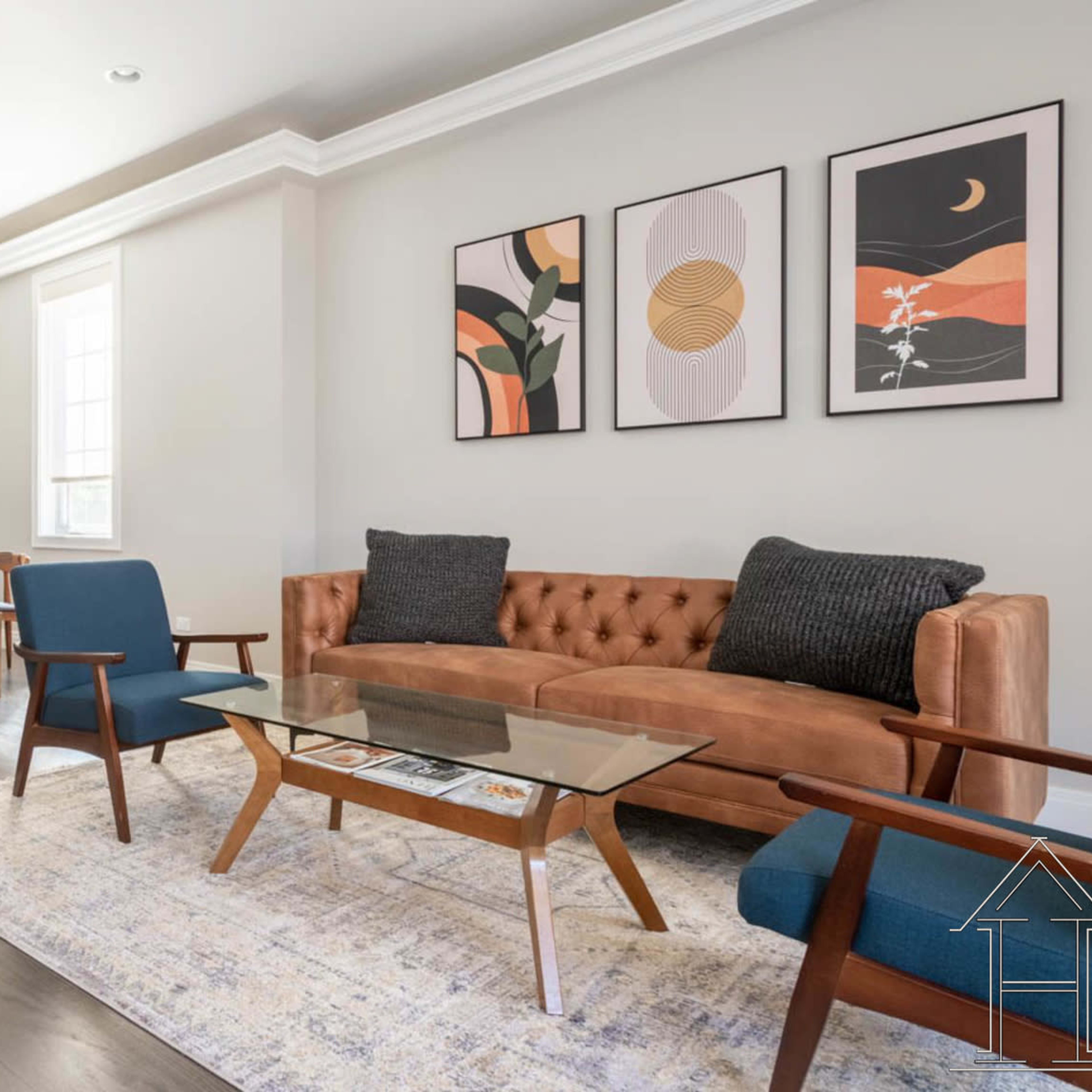 A modern living room with a brown tufted sofa, blue chairs, a glass coffee table, and framed artwork on the walls.
