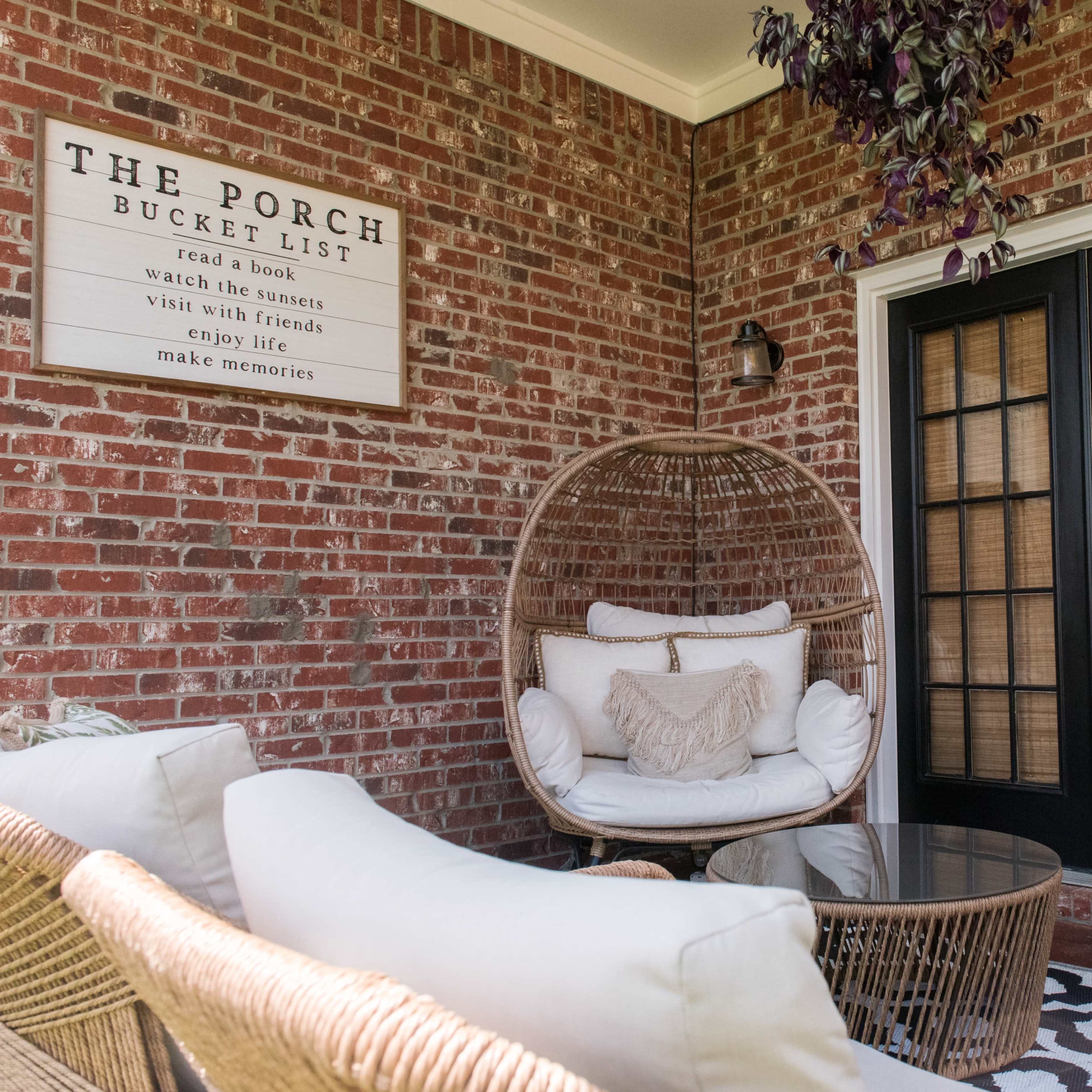 A cozy porch area featuring a hanging rattan chair, a round glass table, and a decorative sign on the brick wall.