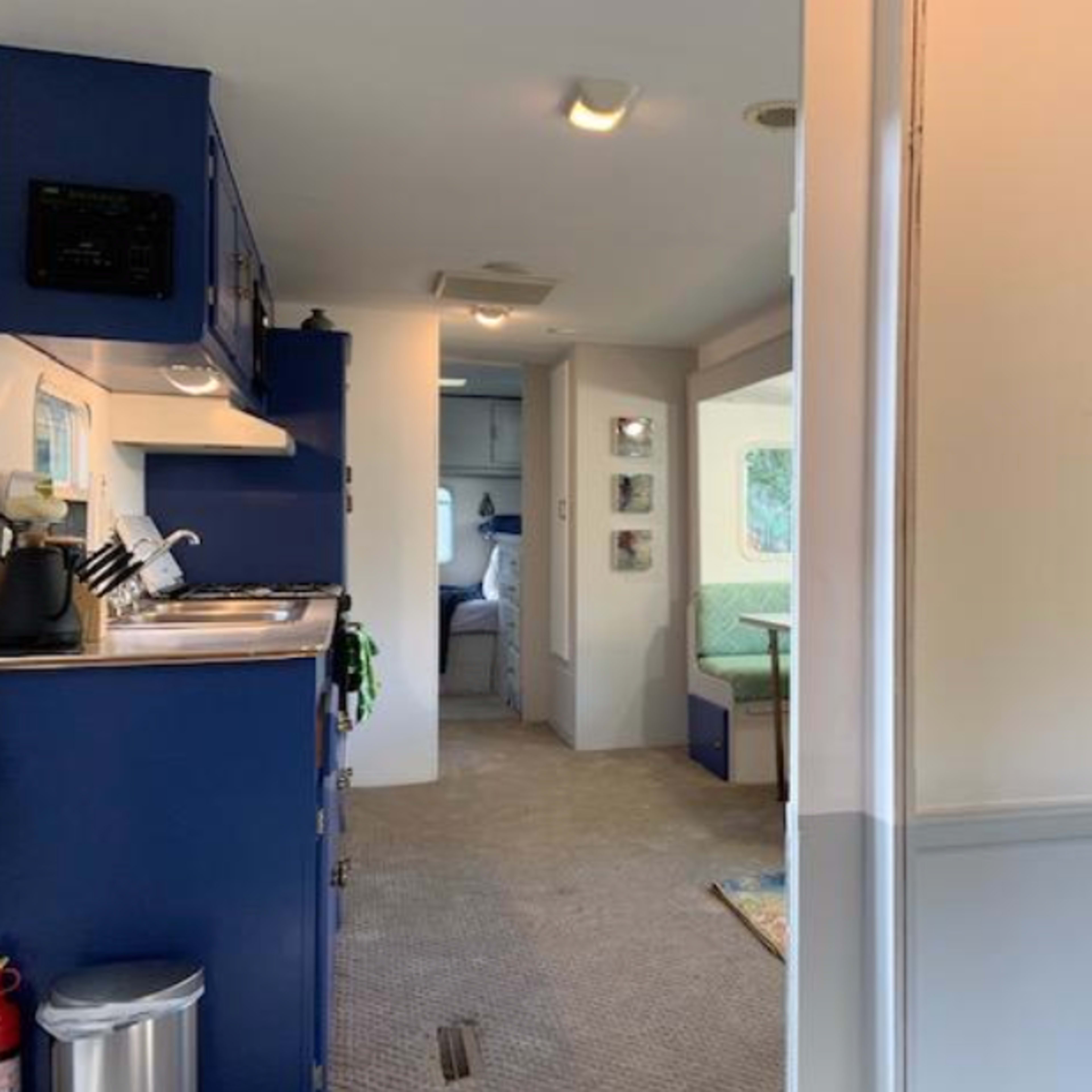 The interior of a camper or mobile home, featuring a kitchen area on the left and a living space further ahead, with a view of a small table and seating area.