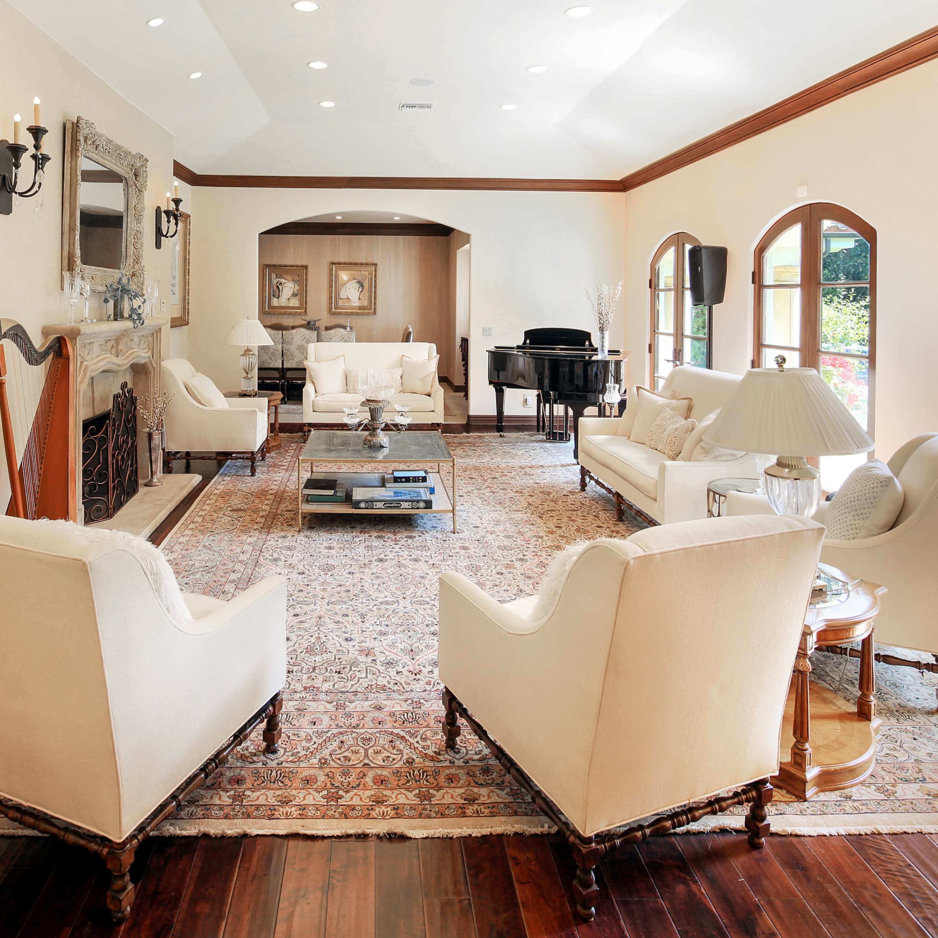 A well-furnished living room with white upholstered chairs, a beige sofa, a large area rug, and a grand piano, all arranged around a central coffee table.