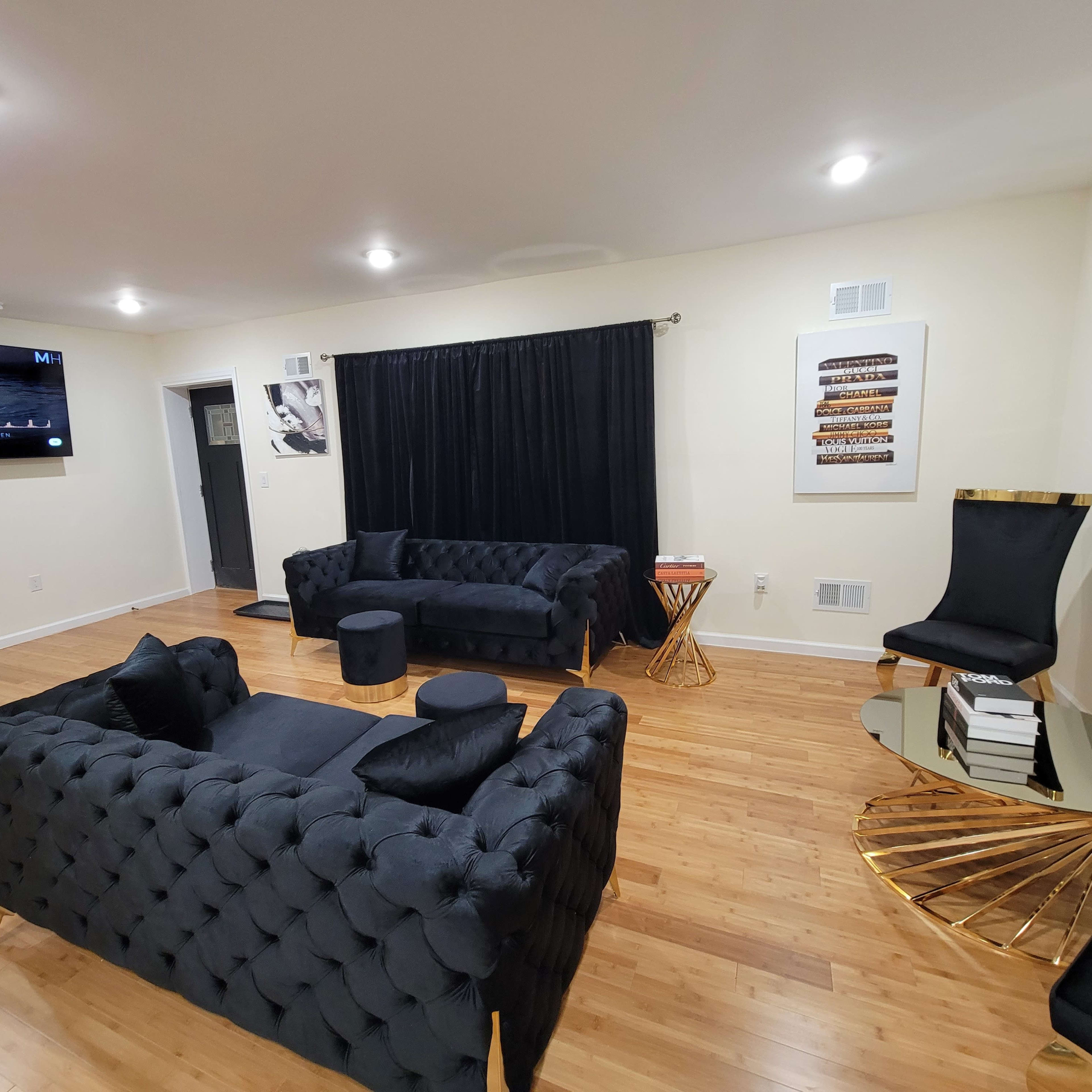 The room features black tufted furniture, a large flat-screen TV on the wall, and a round gold accent table with books.