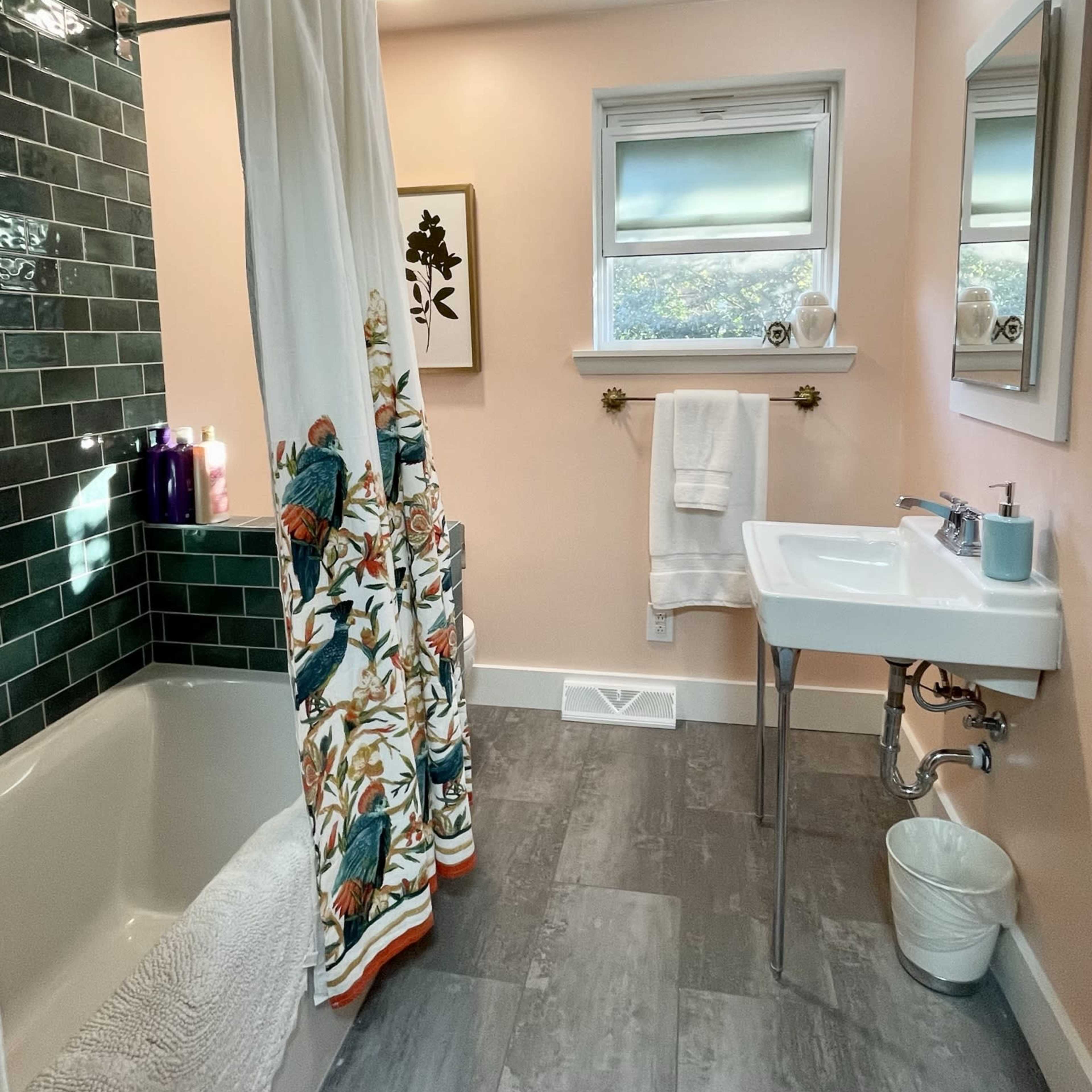 A bathroom features a bathtub with a shower curtain, green tiled wall, white sink on a chrome stand, and soft lighting.