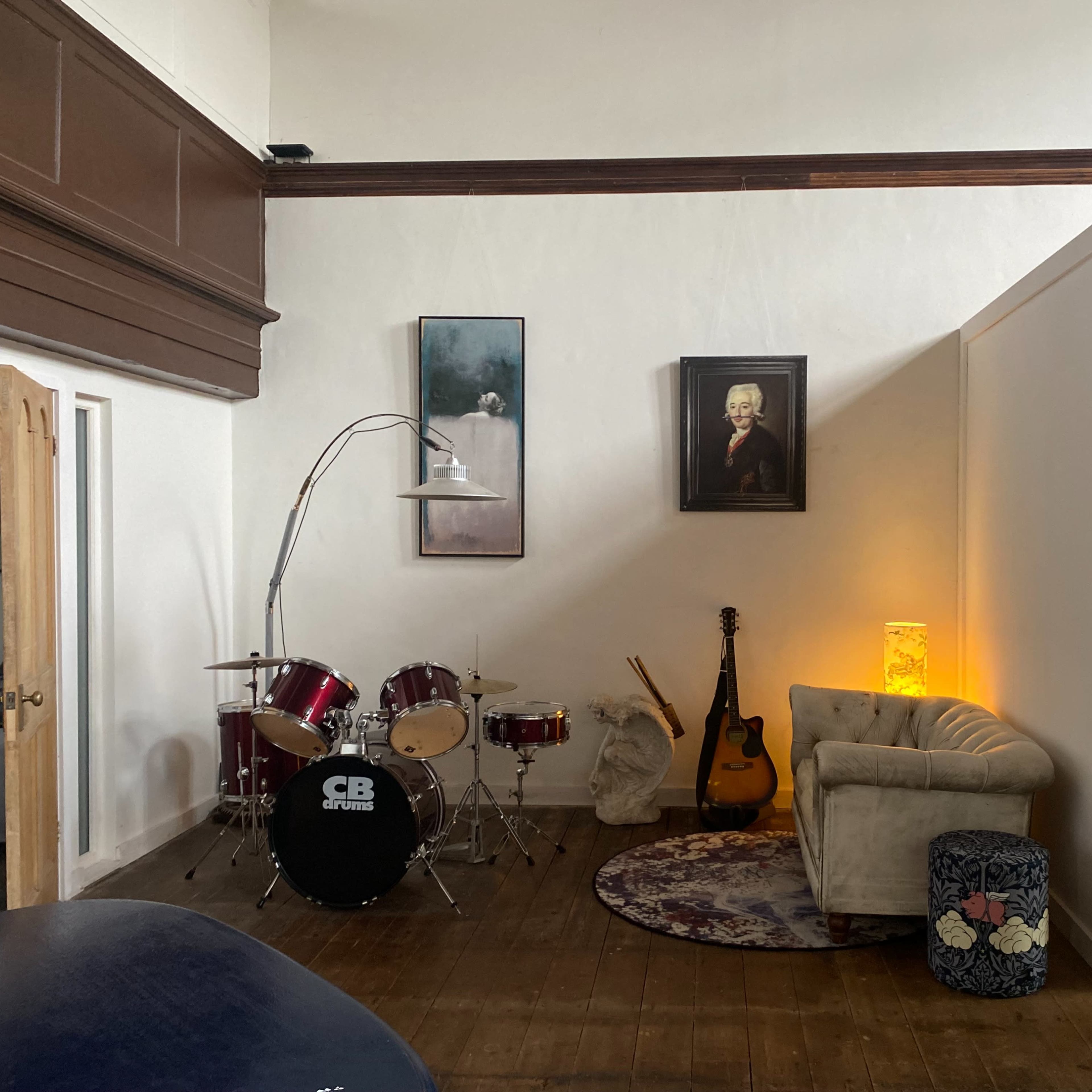 A cozy room with a drum set, an acoustic guitar, and a vintage sofa, illuminated by warm light from a lamp.