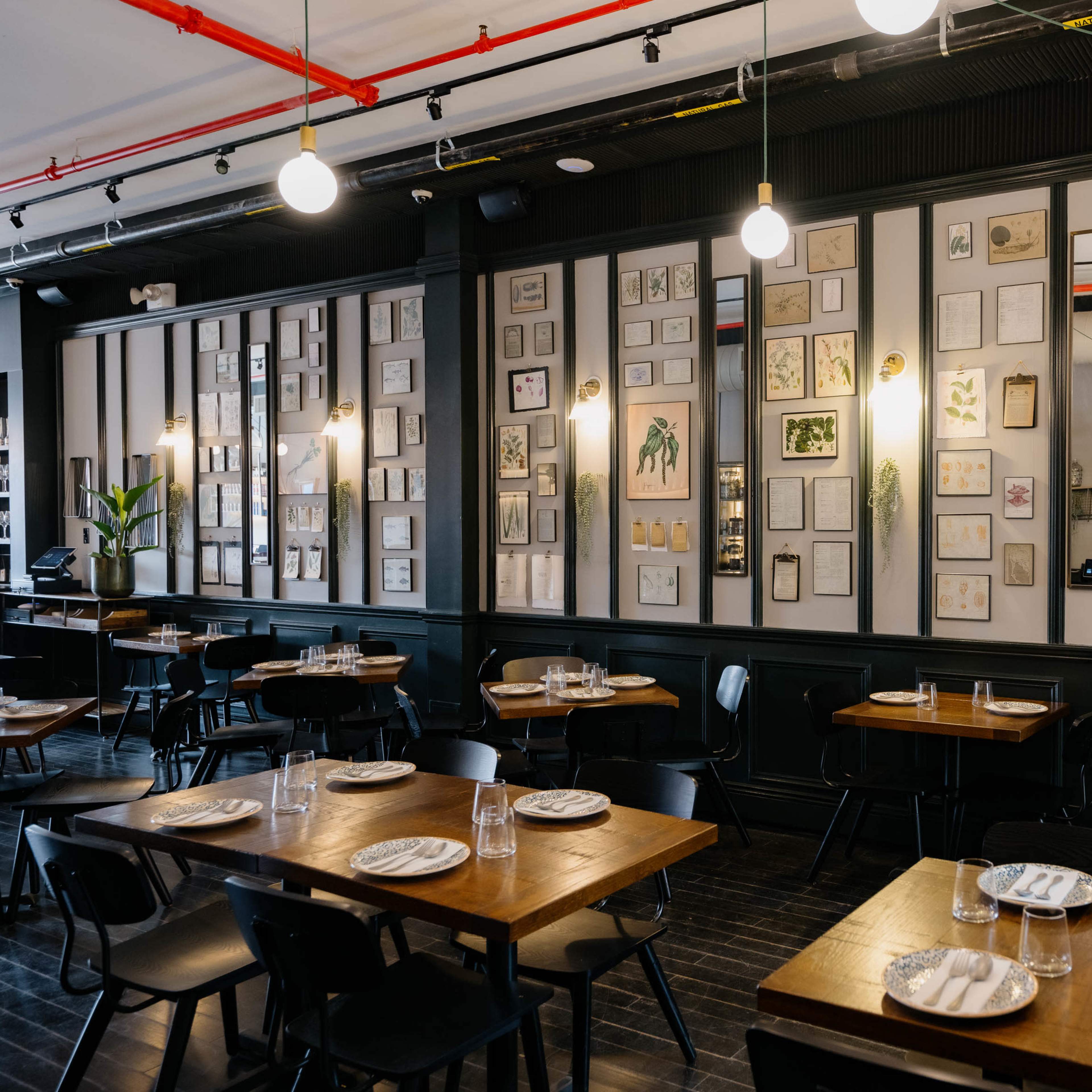 A restaurant interior with wooden tables, black chairs, and decorated walls adorned with various framed artworks and botanical illustrations.