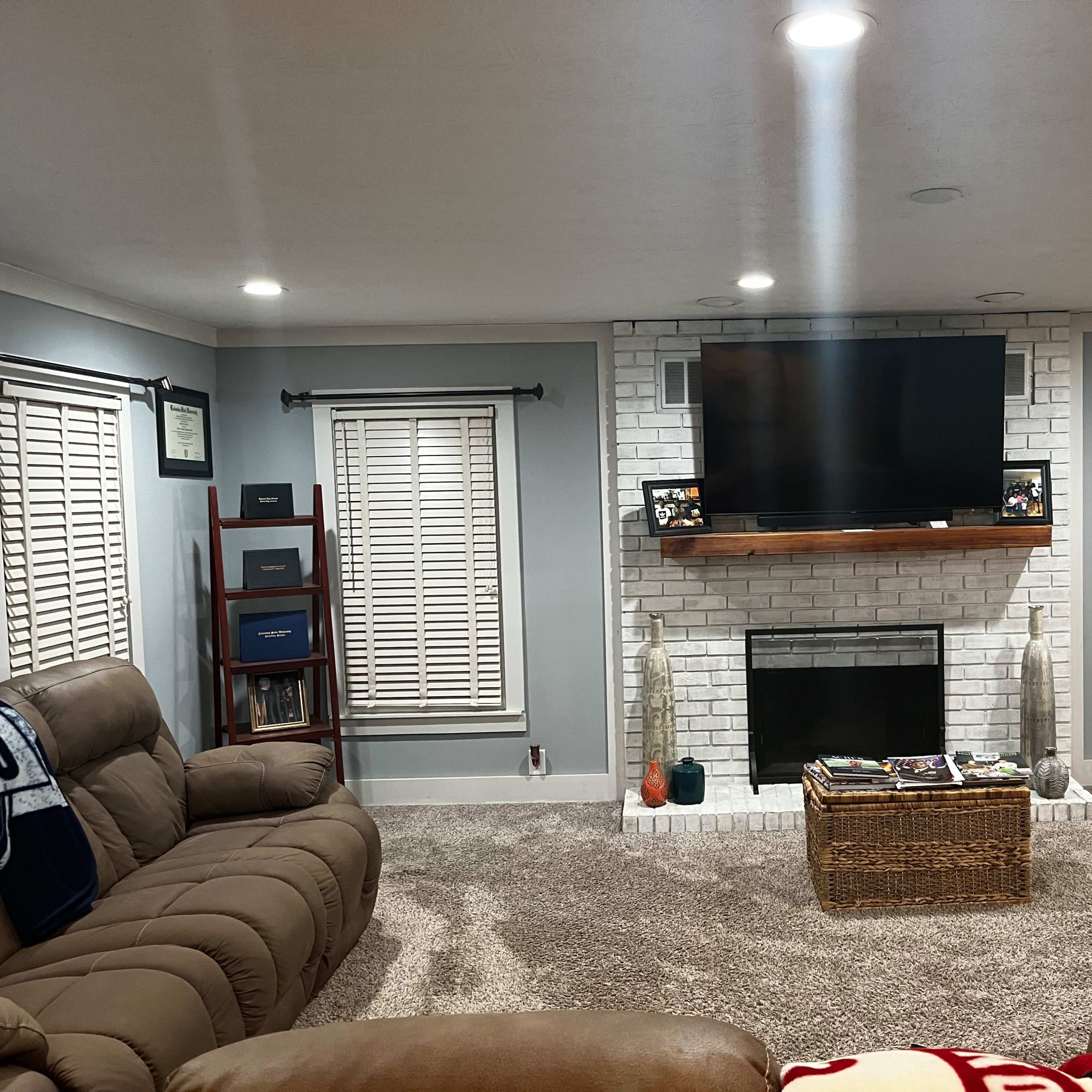 A living room with a light gray color scheme, featuring a white brick fireplace and a mounted television above a wooden shelf.