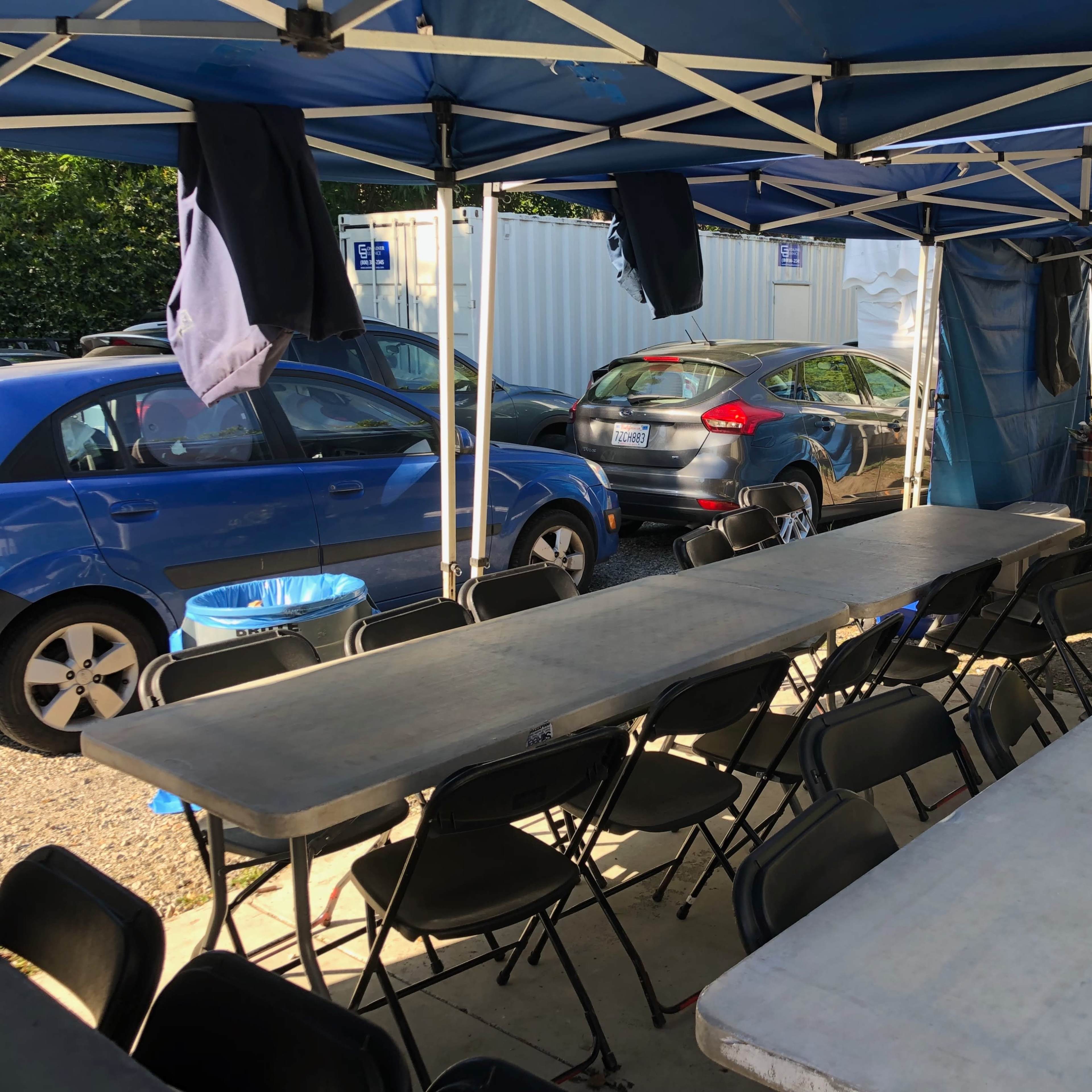 A row of long tables and chairs is set up beneath blue tents, with parked cars visible in the background.