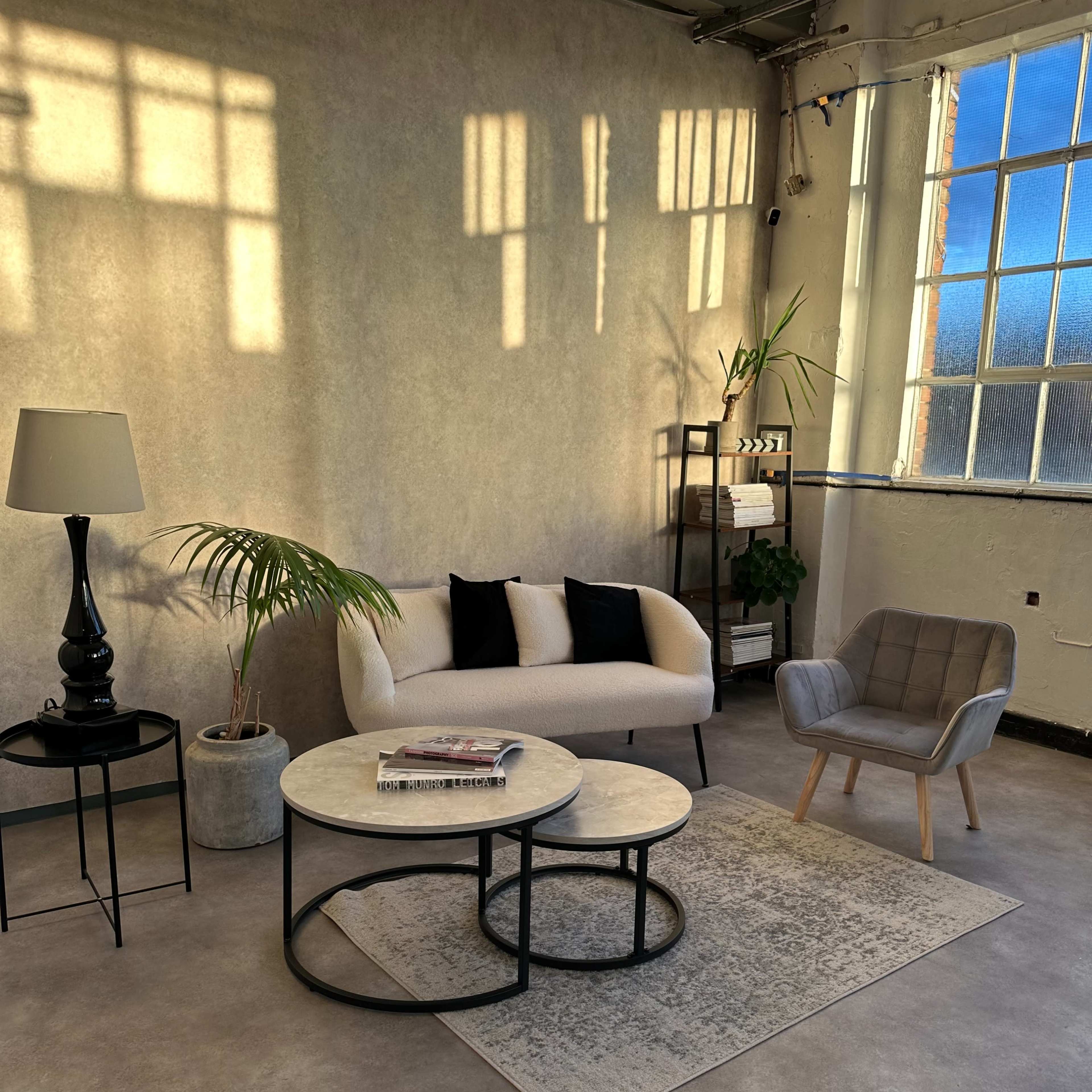 A cozy living area features a beige sofa with black pillows, two round coffee tables, a floor lamp, and a modern armchair, all set against a textured wall and large window.