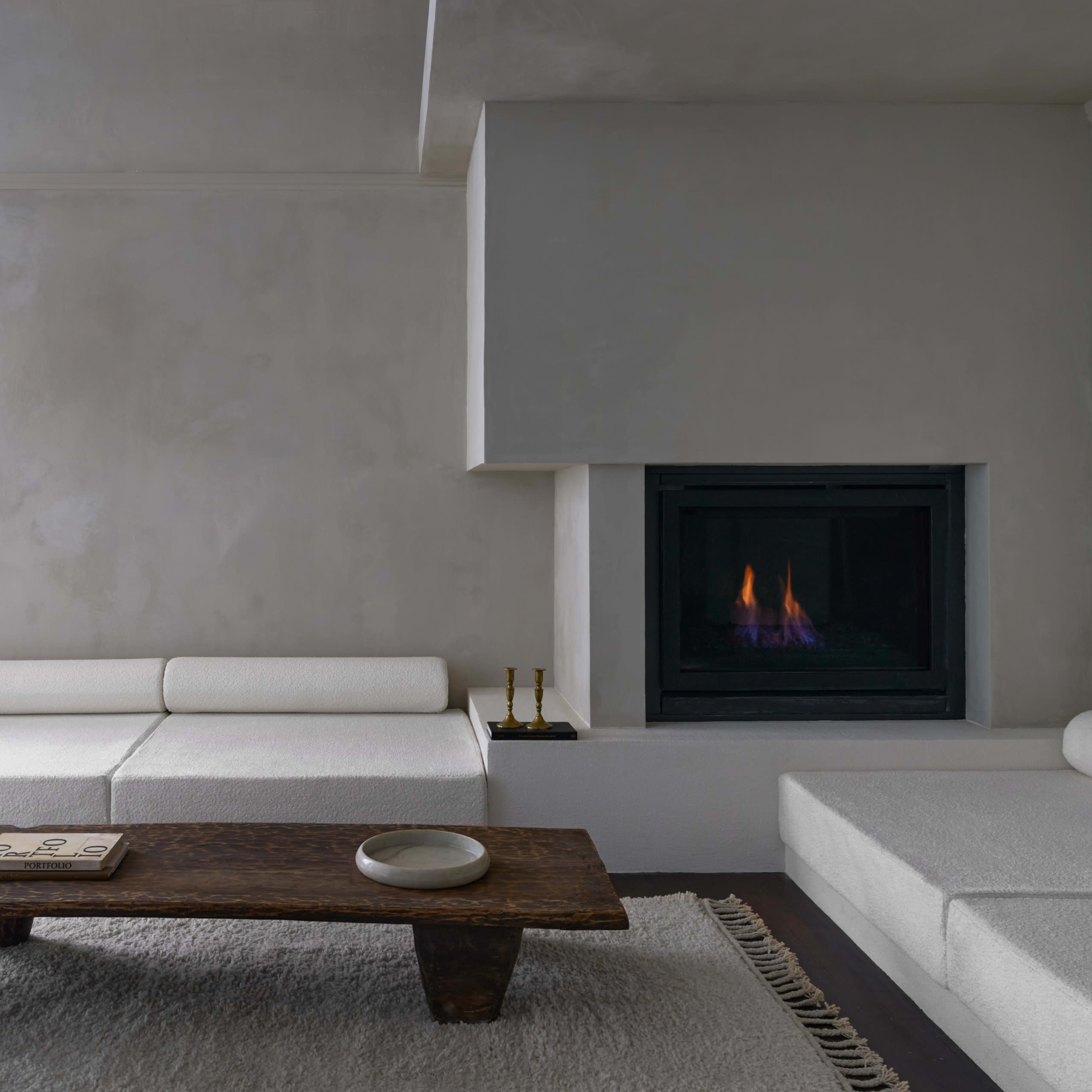 A minimalist living room features a white sectional sofa, a rustic wooden coffee table, and a modern fireplace with a small flame.