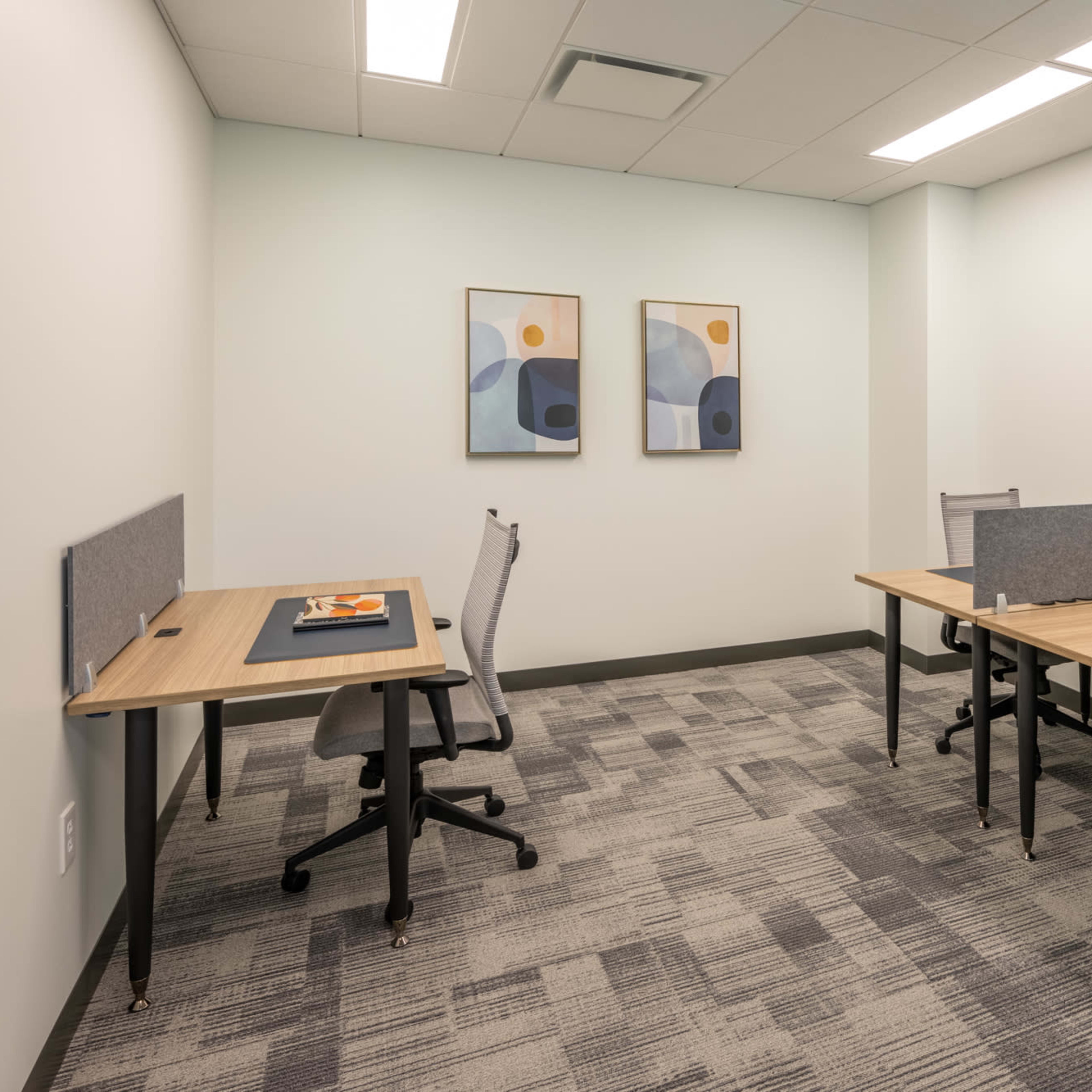 A small office space with two wooden desks, each equipped with a chair and partition, and framed abstract art on the walls.