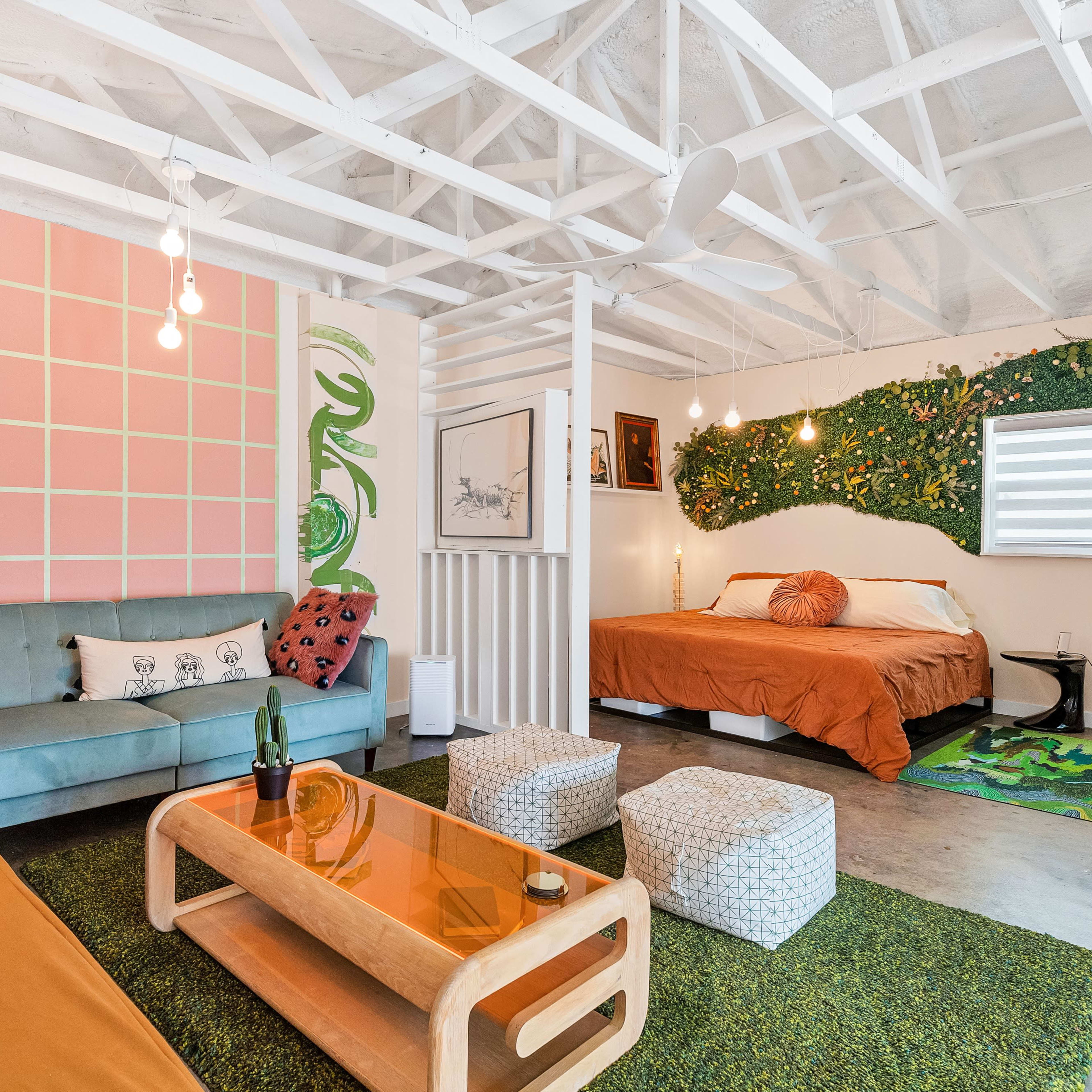 A brightly colored living space featuring a green sofa, a wooden coffee table, a bed with an orange duvet, and decorative plants on the walls.