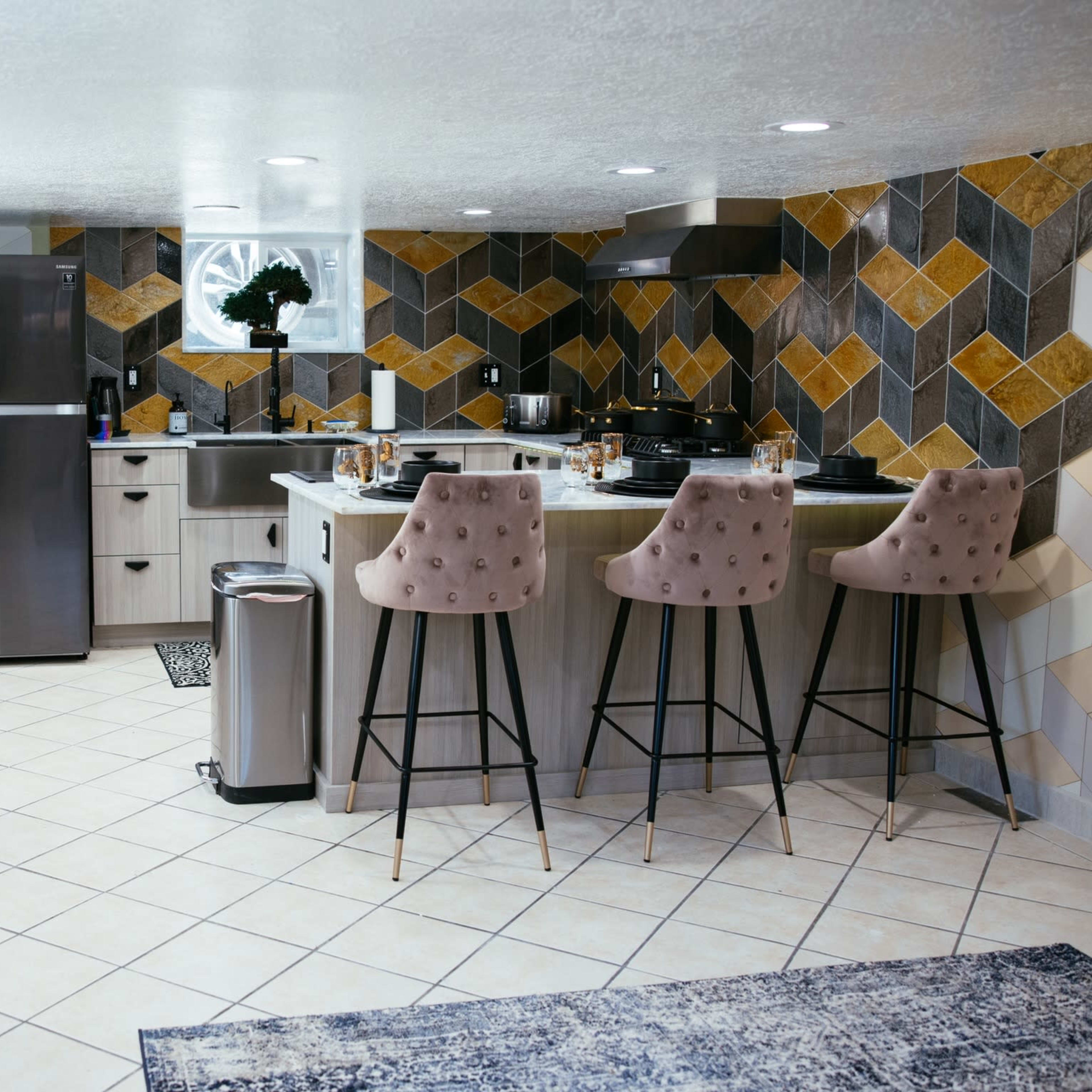 A modern kitchen with a patterned tile backsplash, stainless steel appliances, a dining area with bar stools, and a seating area featuring two chairs.