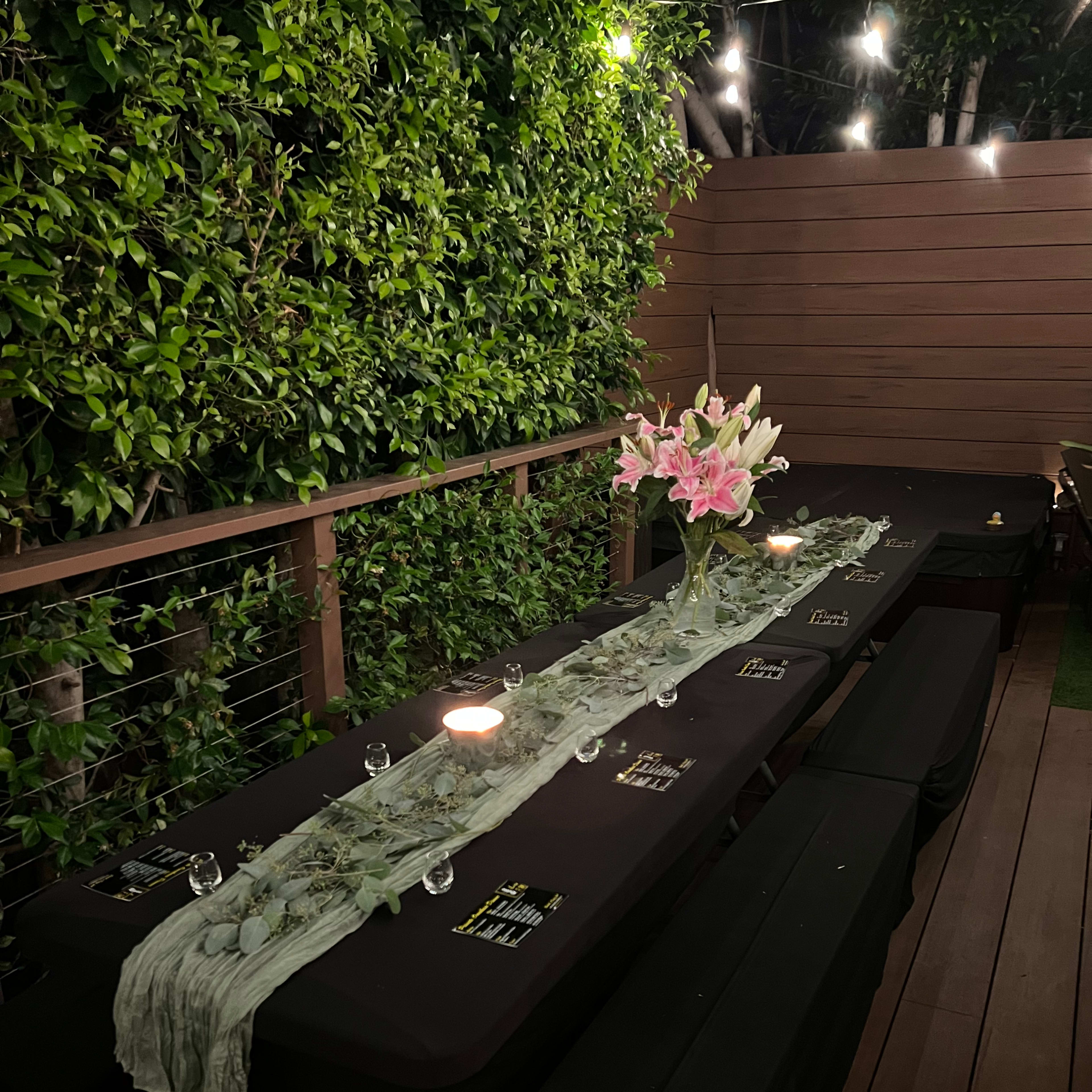 A long dining table with black covers is set outdoors, adorned with a floral centerpiece and surrounded by greenery, illuminated by soft lighting.