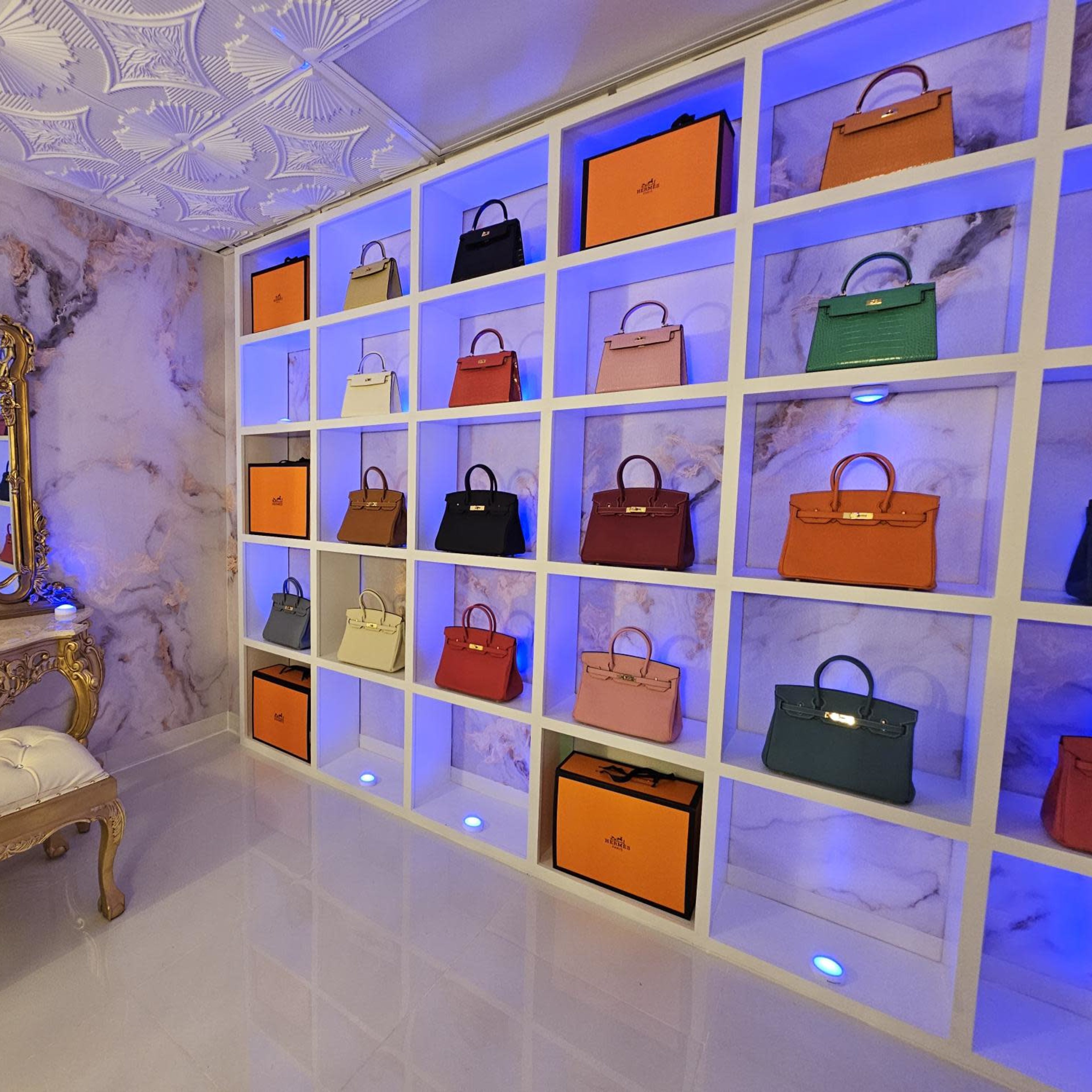 A stylish room with a wall of handbags displayed on shelves, a mirrored ornate vanity, and blue lighting.