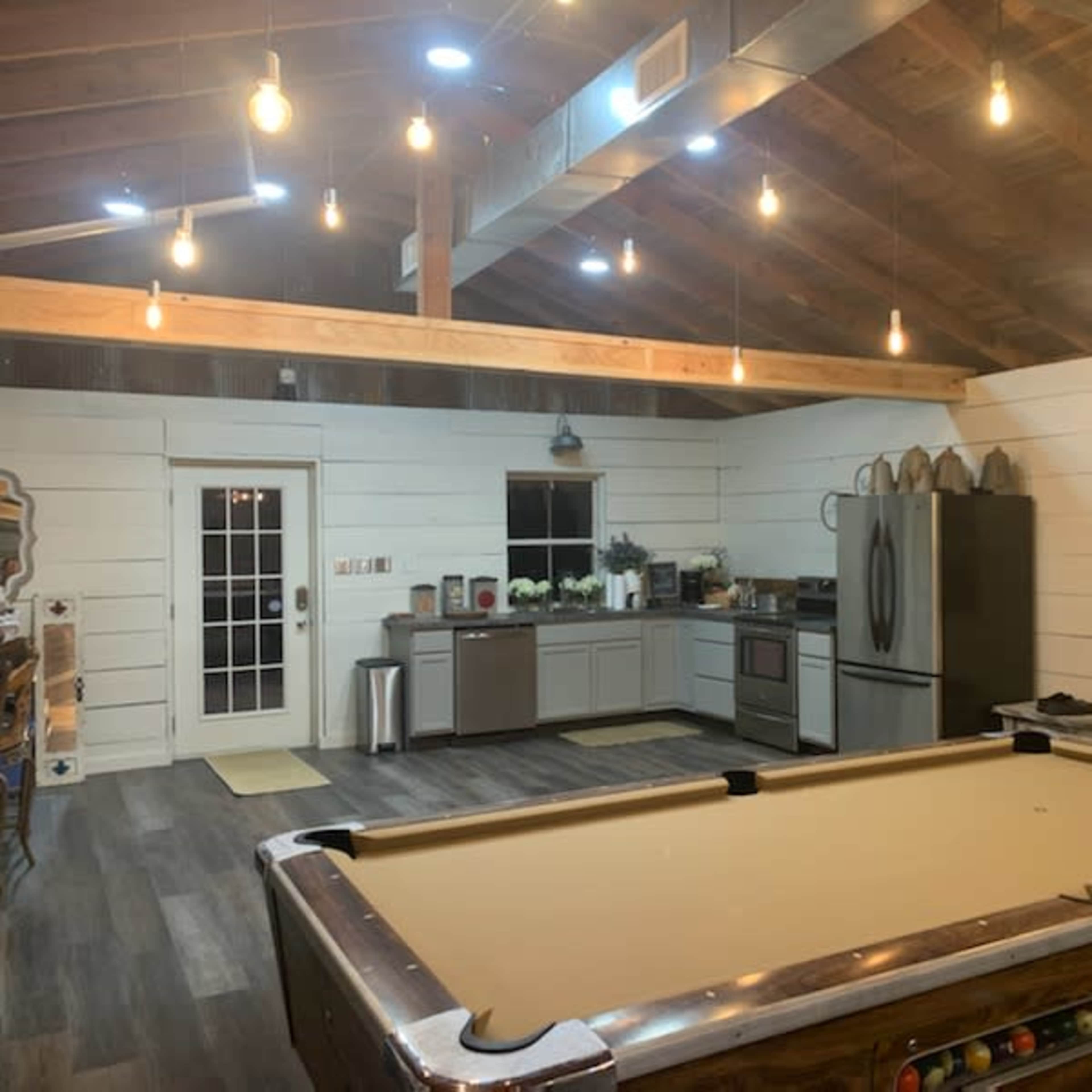 A spacious, well-lit room featuring a pool table, a modern kitchen with stainless steel appliances, and white shiplap walls.