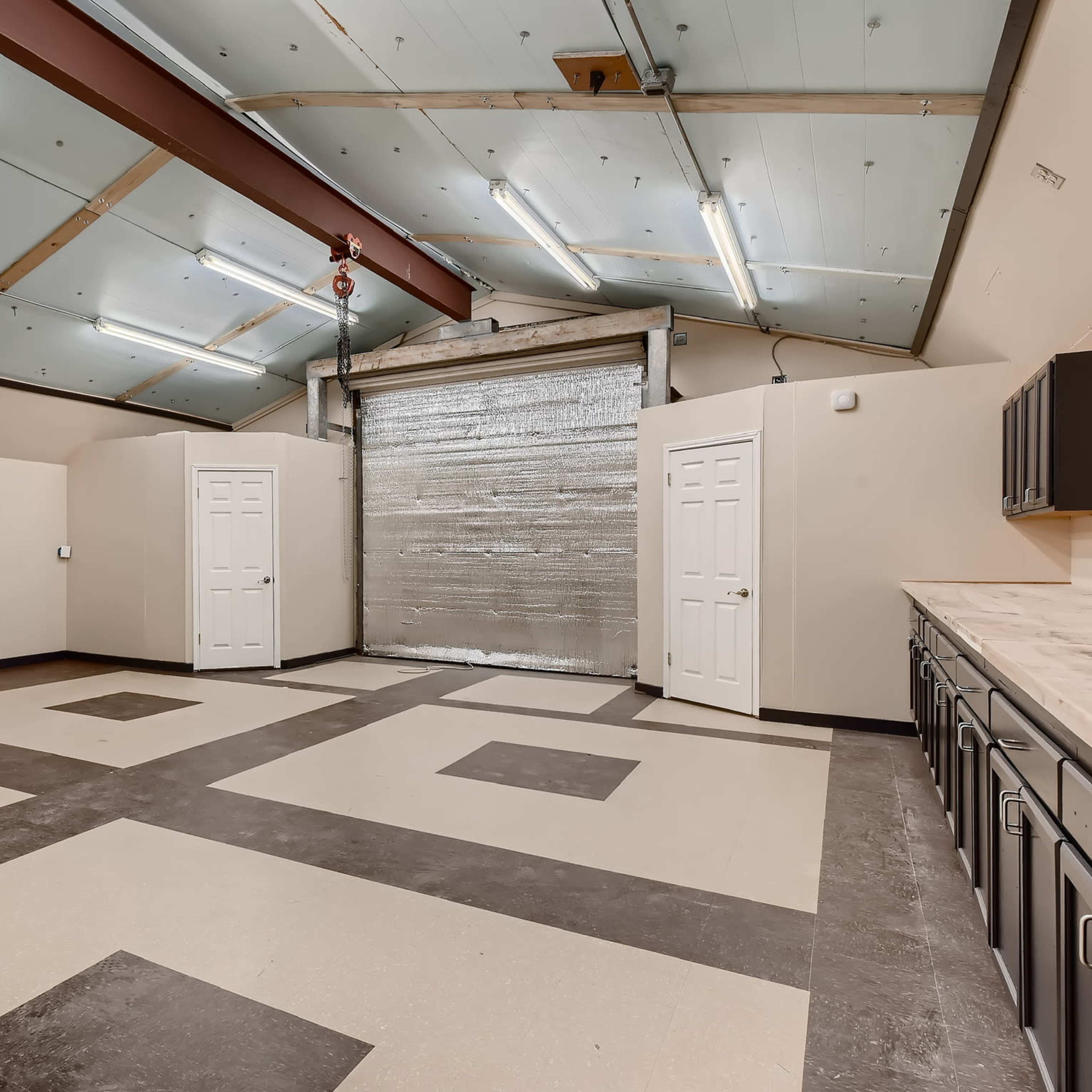 A spacious room with a large roll-up garage door, two white doors, a wall-mounted television, and a tiled floor featuring a geometric pattern.