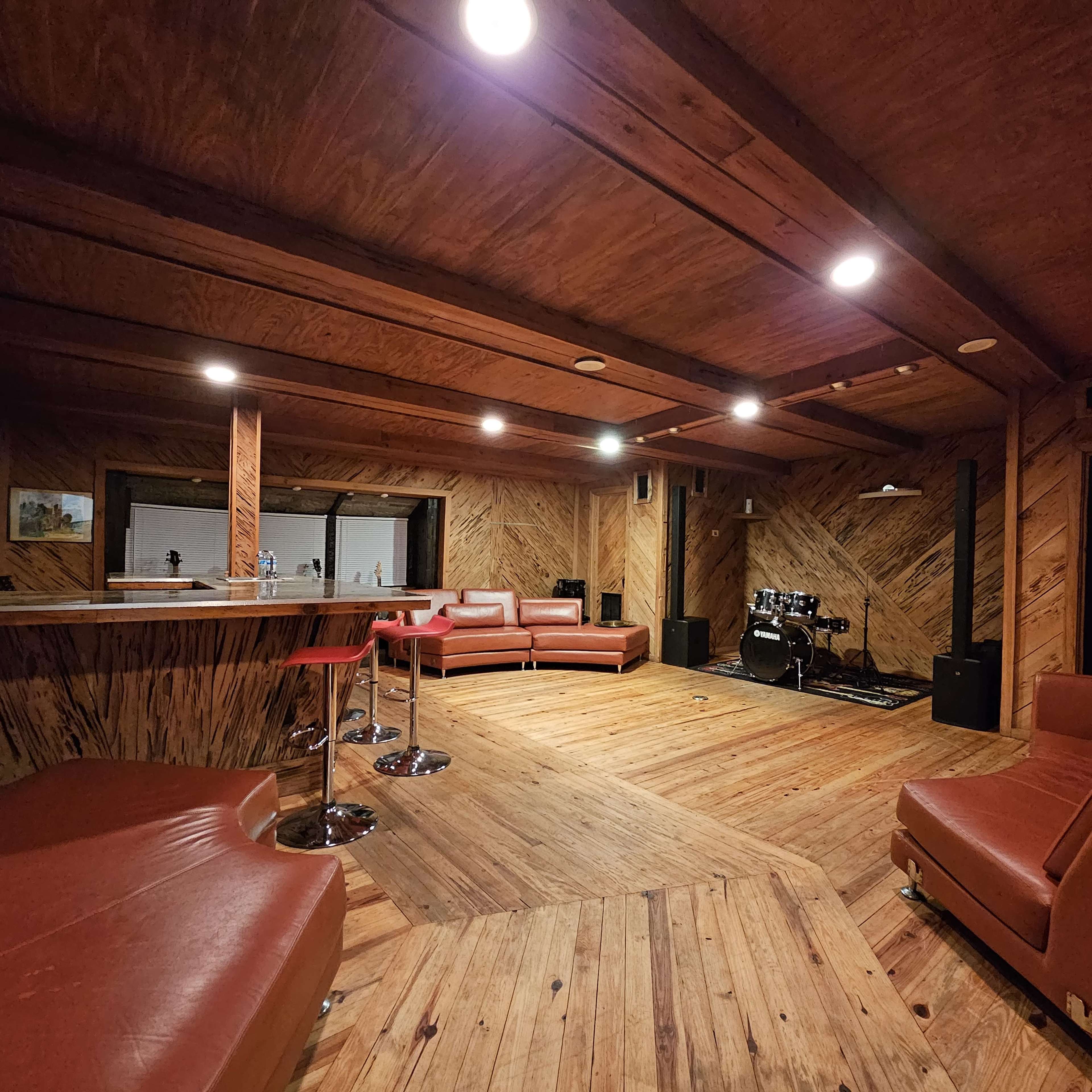 A spacious wooden-paneled room with a bar area, red leather seating, and a small stage equipped with musical instruments.