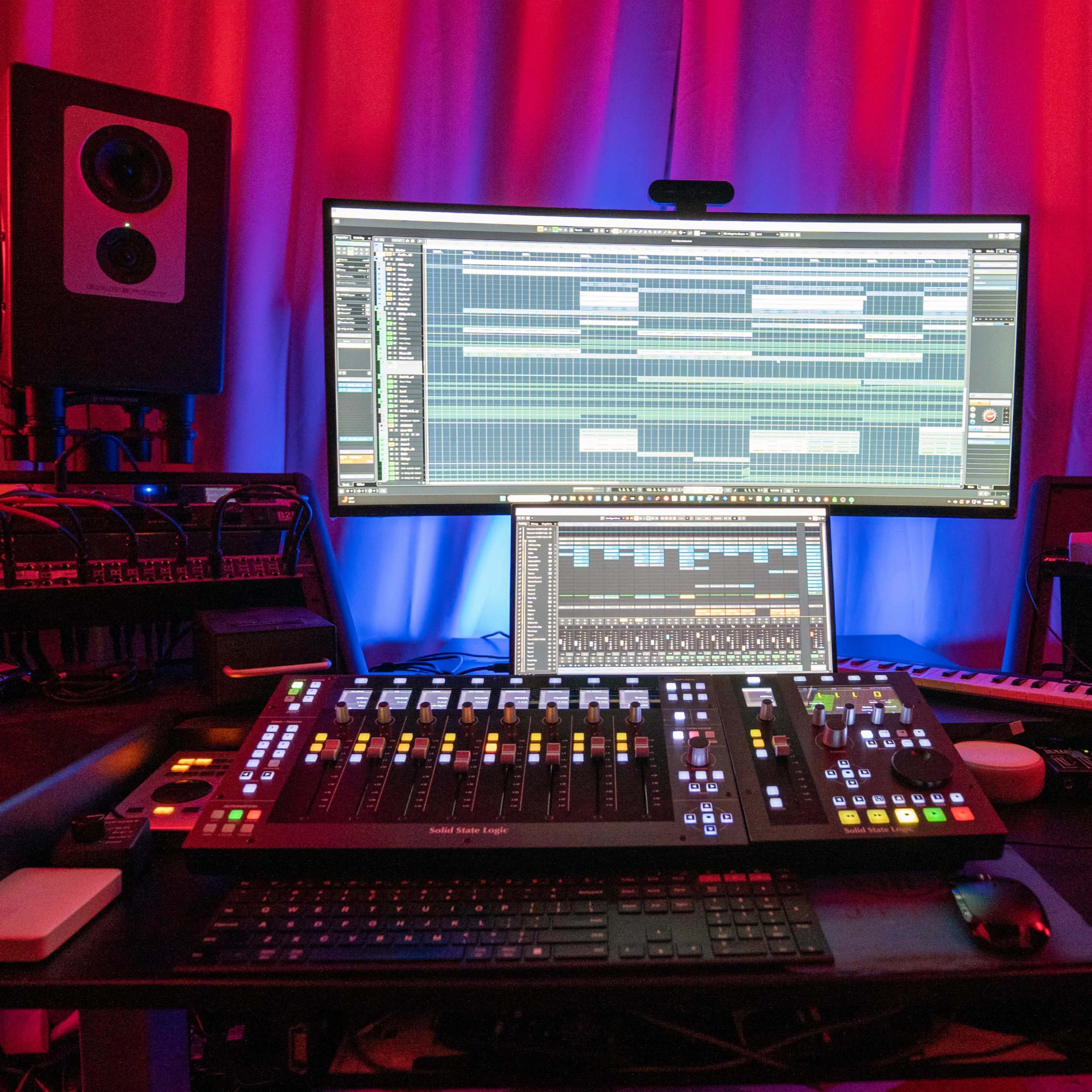 A music production studio featuring a large curved display, multiple audio monitors, and a mixing console with various controls and a computer setup.